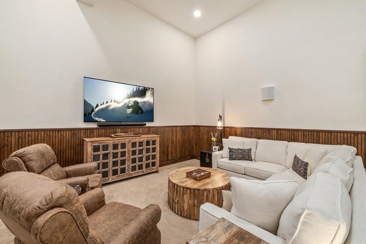 This cozy living room features a warm, rustic aesthetic with wood-paneled wainscoting and a neutral-toned sectional sofa paired with two brown leather armchairs. A large television is mounted on the wall above a wooden media console, while a round, textured wood coffee table sits at the center of the seating area. The room is illuminated by soft overhead lighting, creating an inviting and comfortable space for relaxation.