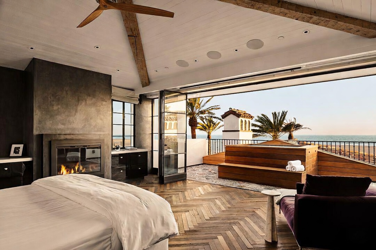 This luxurious primary bedroom features a sophisticated design with a dark, textured fireplace surround and rich herringbone wood flooring. The room seamlessly transitions to an expansive outdoor deck through large glass doors, revealing a private hot tub and panoramic ocean views. The space is accented by a modern ceiling fan and a plush purple lounge chair, creating an elegant and serene coastal retreat.