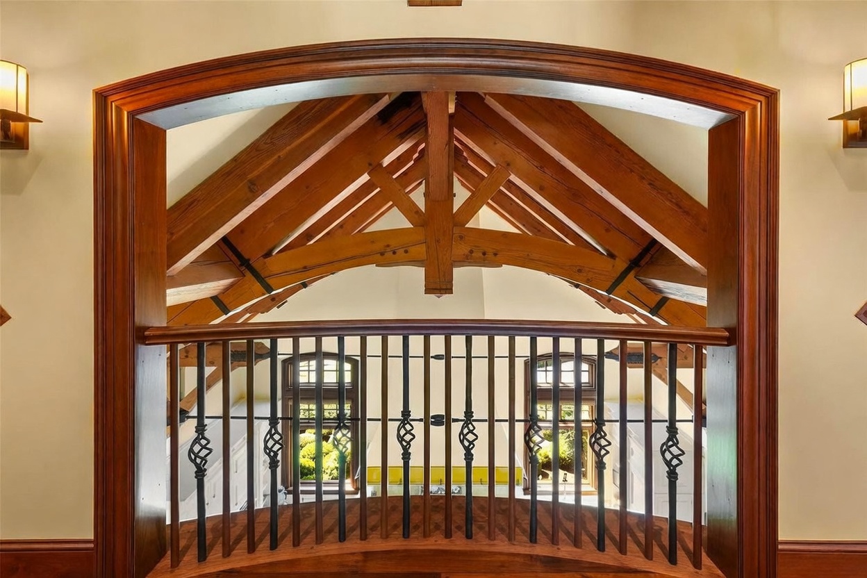 This image captures a view through a prominent, arched wooden doorway that frames a rustic timber-truss ceiling structure. A decorative metal railing with twisted balusters separates the foreground from the open space beyond, which features large windows looking out onto greenery. The perspective is centered and elevated, emphasizing the craftsmanship of the exposed wood beams and the elegant transition between interior spaces.