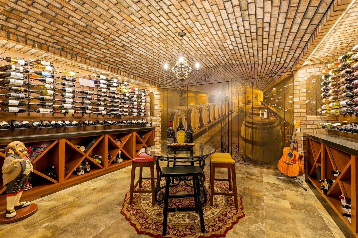 This sophisticated wine cellar features a distinctive barrel-vaulted brick ceiling and a mural depicting a wine cave, creating an immersive atmosphere. The space is equipped with extensive wall-mounted wine racks, a central bistro-style table with stools, and a decorative figurine, all set upon a tiled floor. A guitar stands in the corner, adding a personal touch to this well-appointed entertainment area.