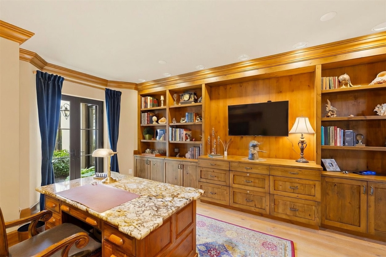This sophisticated home office features extensive custom wood cabinetry with integrated shelving and a central television mount. A large desk with a stone top sits in the foreground, complemented by a patterned area rug and a classic banker's lamp. French doors with dark blue curtains provide natural light and access to the outdoors, creating a professional yet inviting workspace.