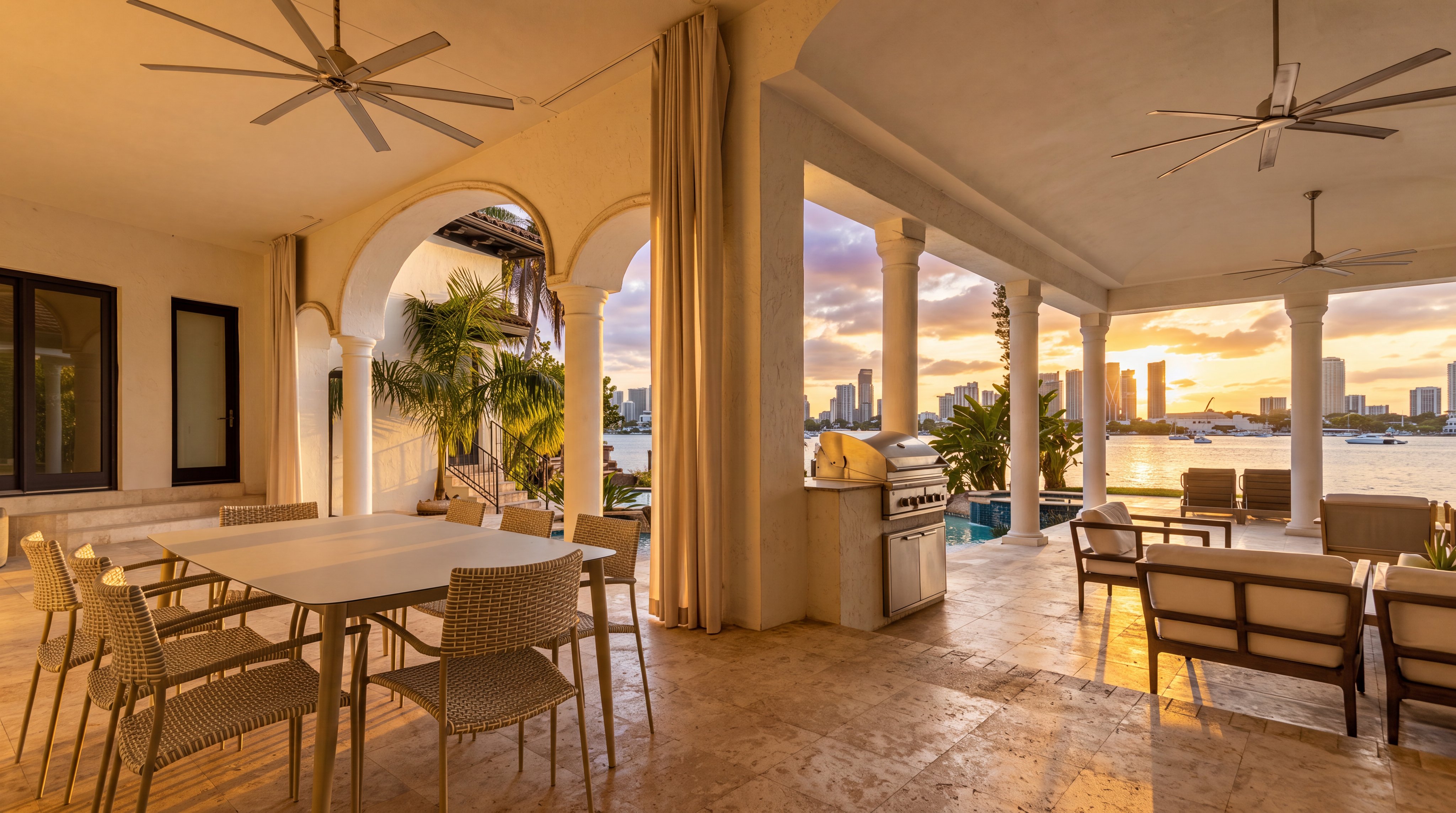 This elegant covered outdoor patio features a spacious dining area, a built-in outdoor kitchen, and comfortable lounge seating, making it perfect for refined waterfront entertaining. The living space is defined by classical arched columns, natural stone flooring, and expansive views of the bay and city skyline at sunset. It offers a luxurious, resort-style atmosphere with seamless indoor-outdoor connectivity.