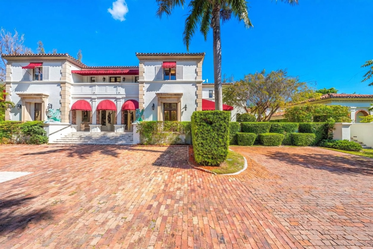 This grand Mediterranean-style estate features a symmetrical white stucco facade accented by striking red awnings and decorative stone detailing. A wide, circular brick-paved driveway leads to a prominent central entrance with stone steps, flanked by manicured hedges and lush landscaping. The scene is captured from a low-angle perspective, emphasizing the imposing scale and architectural elegance of the residence under a clear blue sky.