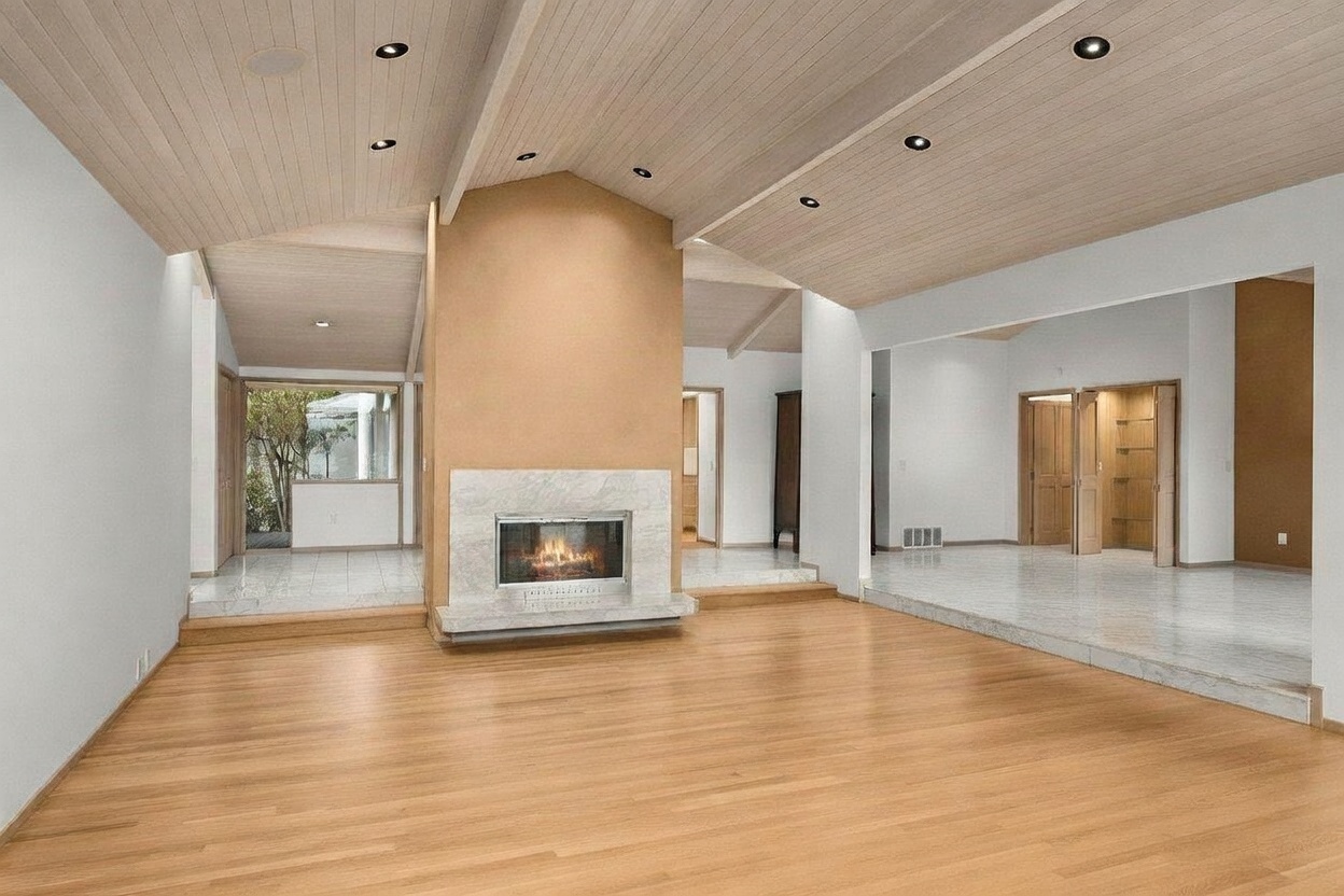 This spacious living area features a prominent central fireplace with a light-colored stone surround and a hearth that separates the room into distinct zones. The space is defined by light wood flooring in the main area and elevated stone-tiled platforms on either side, all beneath a vaulted, light-toned wood-paneled ceiling. The open-concept layout creates a bright and airy atmosphere, with recessed lighting and a view through to an adjacent room with large windows.