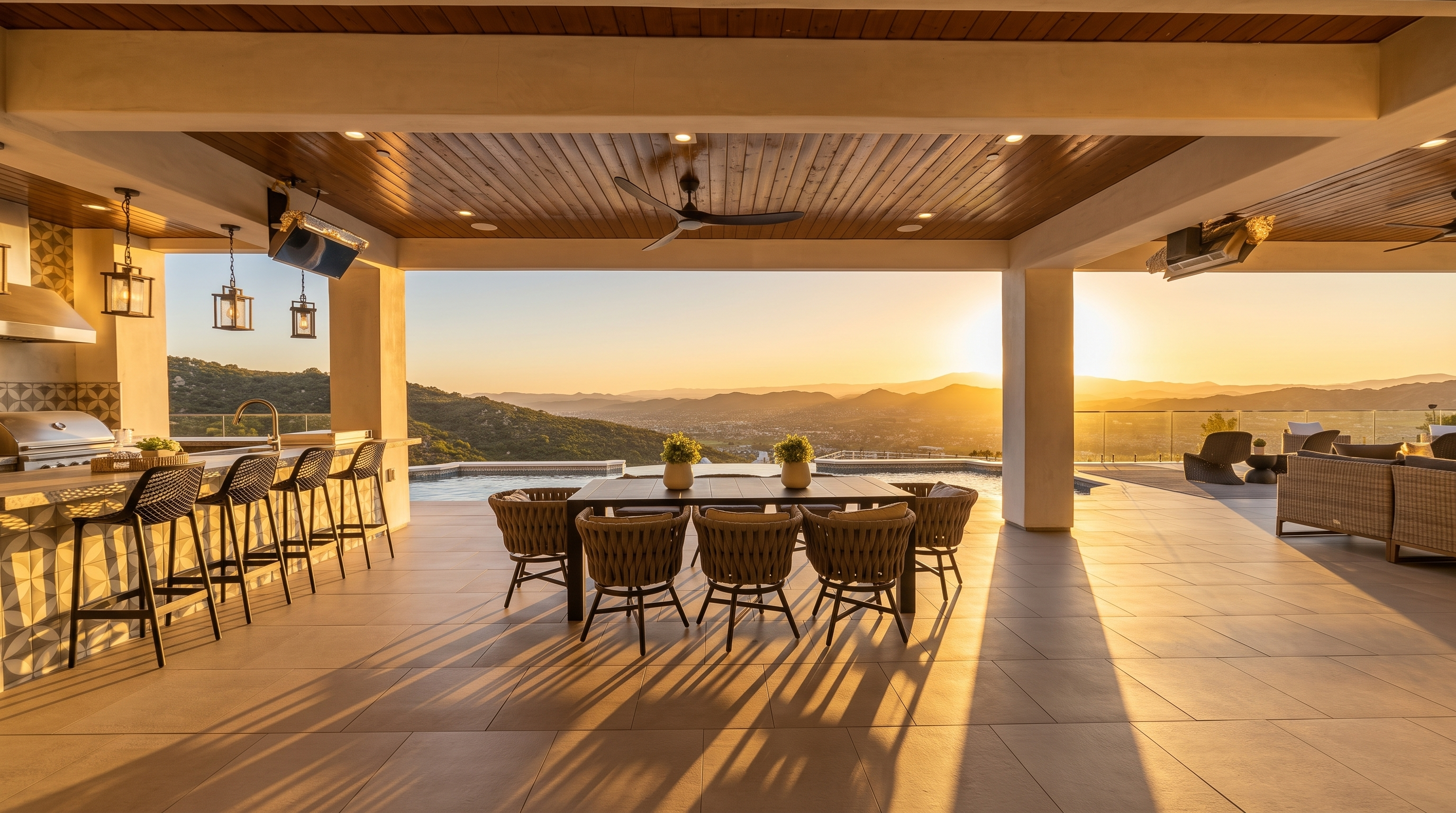 This luxurious covered outdoor living space seamlessly blends indoor comfort with an expansive panoramic view of the surrounding hills and valley. The area features a full bar with stylish wicker stools and a spacious dining set, all under a beautiful wood-paneled ceiling with modern recessed lighting and a ceiling fan. The seamless transition to a stunning infinity-edge pool highlights the high-end, resort-style appeal of this property.