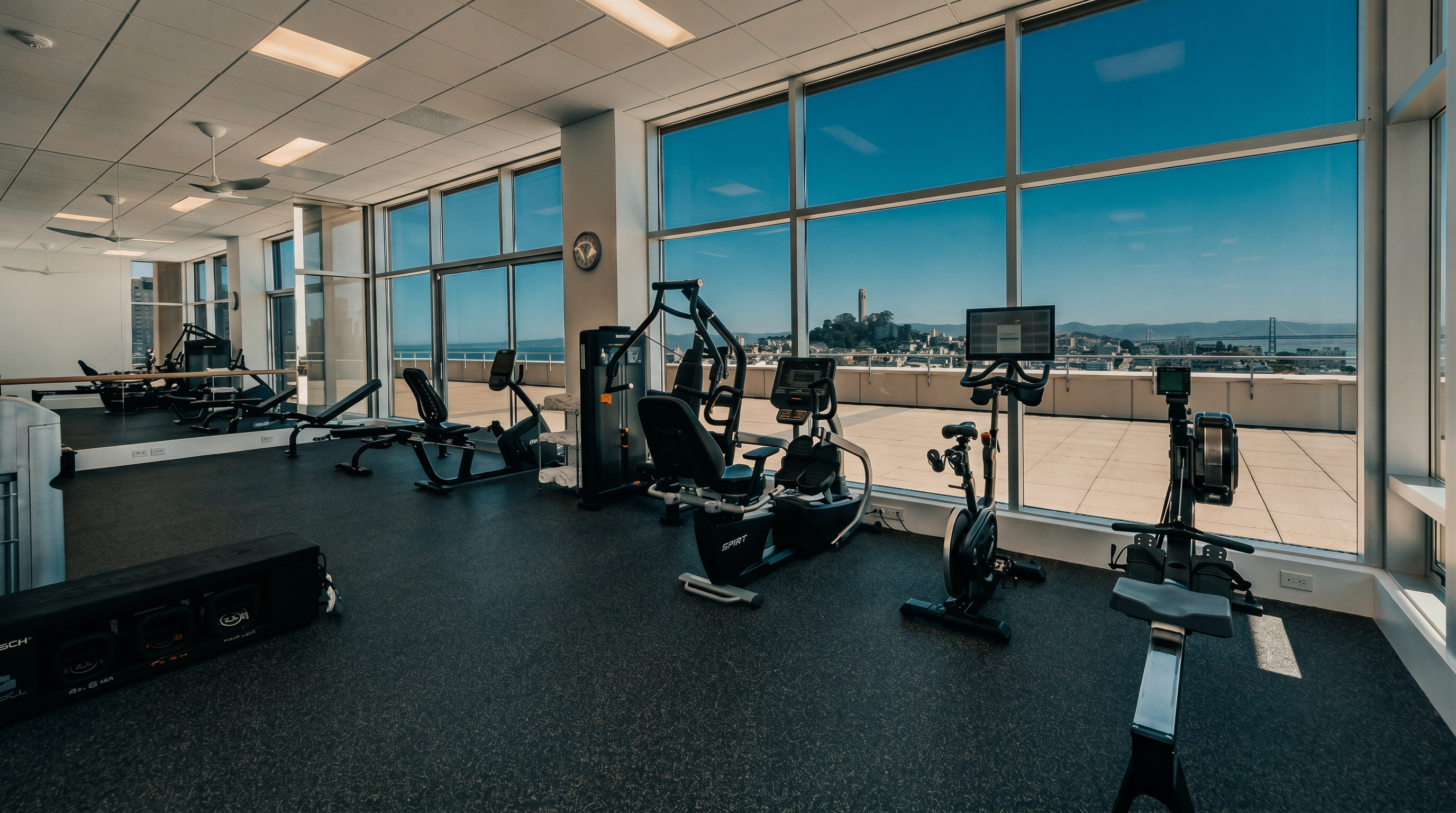 This bright and spacious home gym features a variety of professional-grade exercise equipment, including a rowing machine, stationary bike, and strength training stations. The room is defined by expansive floor-to-ceiling windows that provide an unobstructed, panoramic view of the city skyline and bay. The space is finished with durable, dark rubberized flooring, creating a clean and functional environment for fitness.