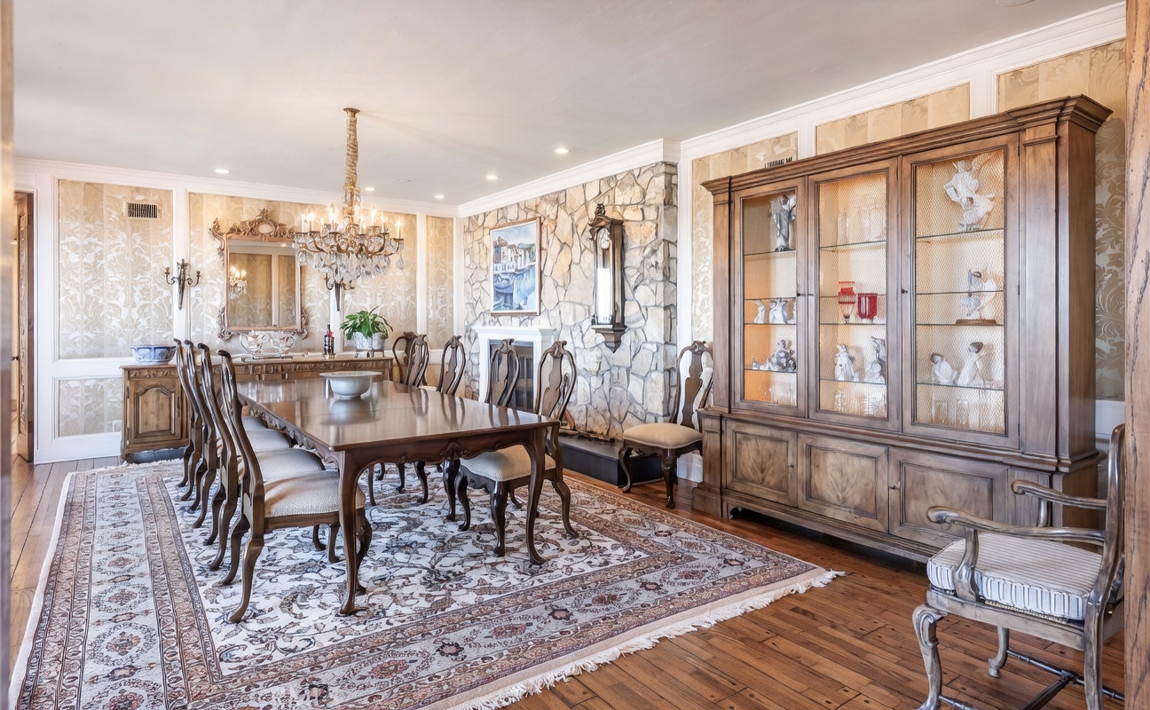 This formal dining room features a large wooden table surrounded by upholstered chairs, set upon an intricate patterned area rug. The space is anchored by a stone-clad chimney breast and a substantial wooden hutch with glass-front cabinets displaying decorative items. A crystal chandelier hangs above the table, while the walls are adorned with patterned wallpaper and wainscoting, creating a traditional and elegant atmosphere.