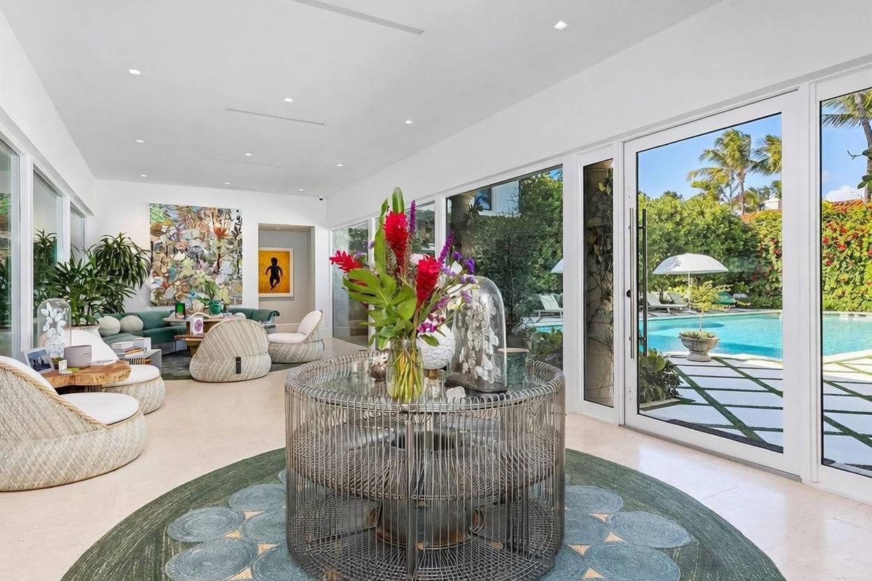 This bright and airy living space features a seamless transition to the outdoors through expansive floor-to-ceiling glass doors that overlook a private swimming pool. The room is furnished with contemporary, textured seating and a central circular table topped with a vibrant floral arrangement, all set upon a patterned area rug. The interior design emphasizes a sophisticated, open-concept aesthetic with neutral tones and high-end artistic accents.