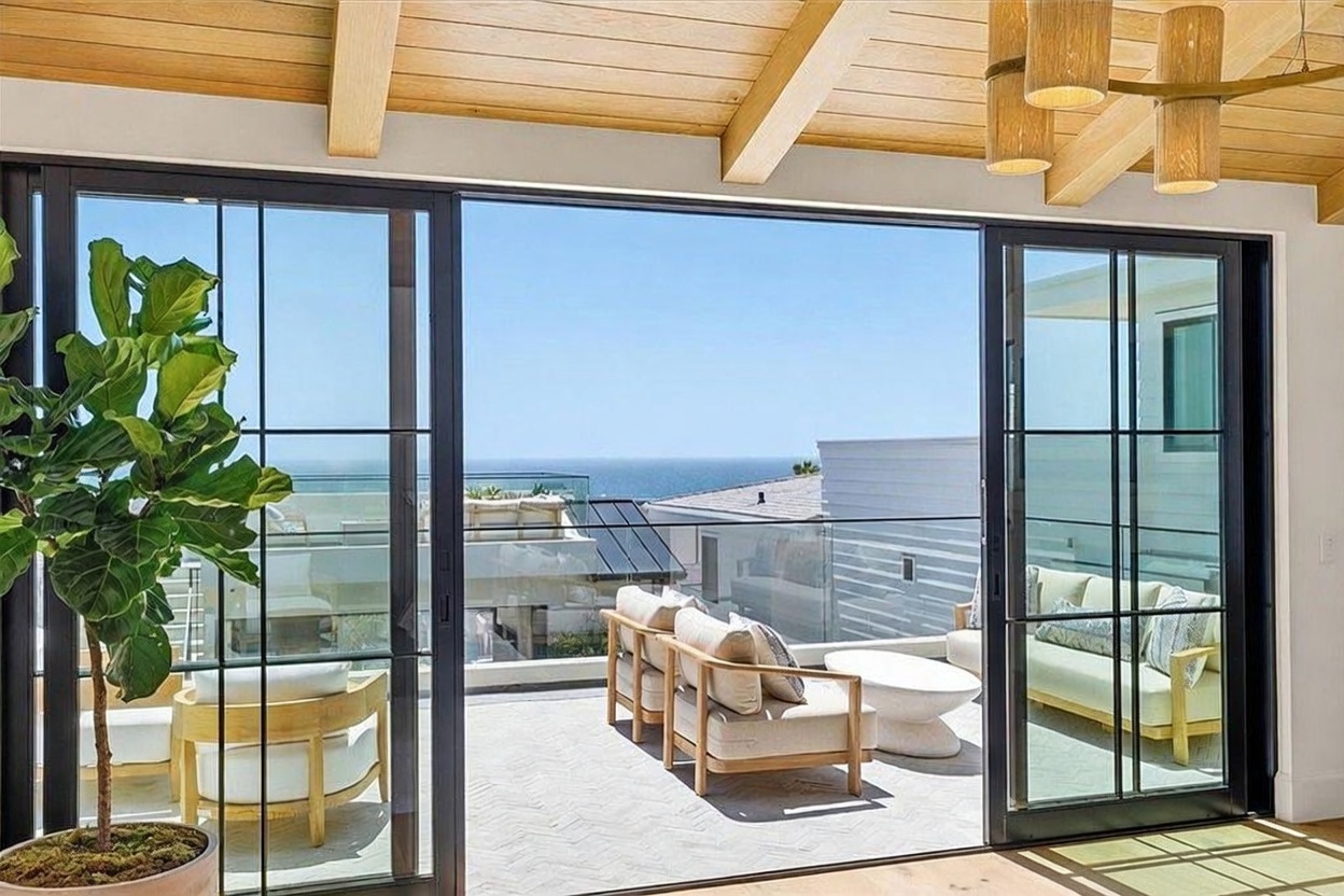 This interior space features expansive floor-to-ceiling sliding glass doors that open onto a private balcony with ocean views. The room is accented by a light wood-paneled ceiling and a modern, multi-tiered pendant light fixture, while a large potted plant adds a touch of greenery to the foreground. The balcony is furnished with comfortable outdoor seating, creating a seamless transition between indoor living and the coastal environment.