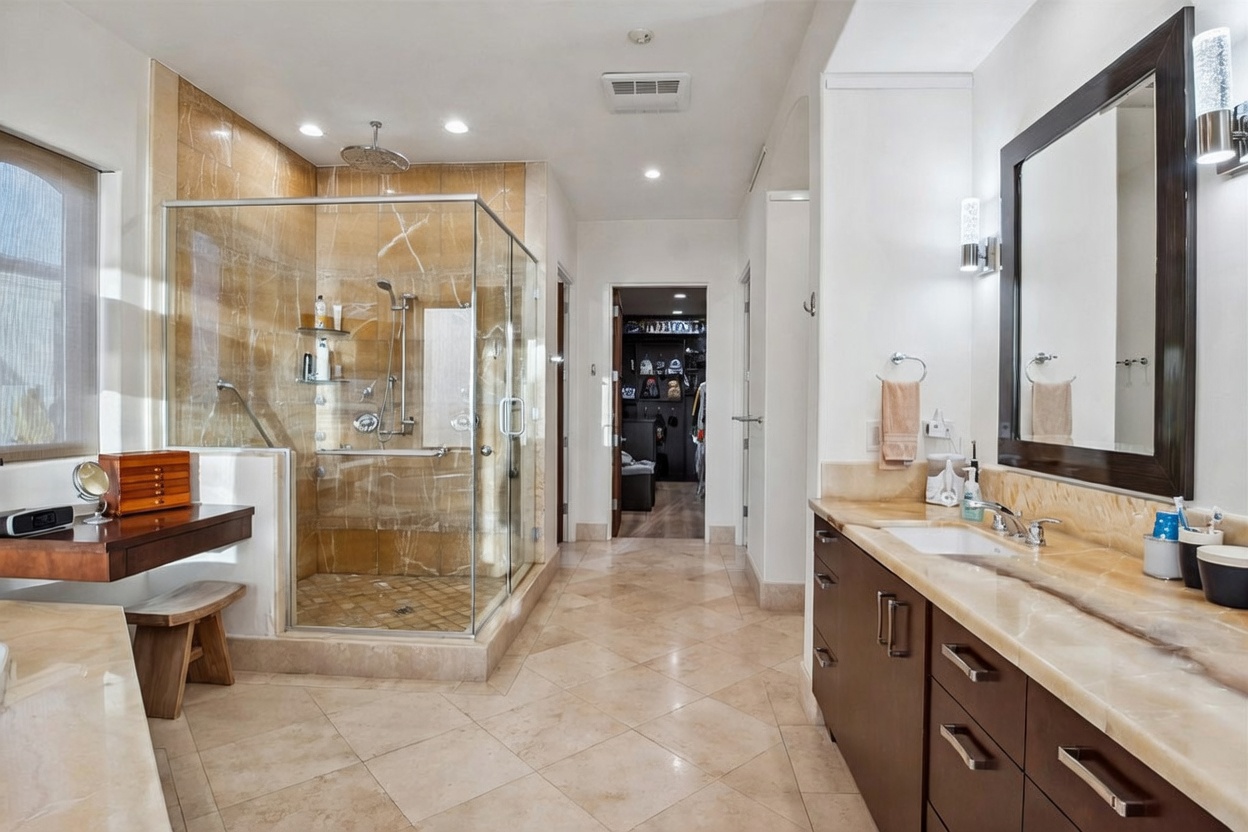 This spacious primary bathroom features a large glass-enclosed shower with stone-tiled walls and a built-in bench. To the right, a long vanity with dark cabinetry and a stone countertop provides ample storage, while a built-in vanity desk with a wooden stool sits to the left. The room is finished with neutral-toned floor tiles and leads directly into a walk-in closet at the far end.