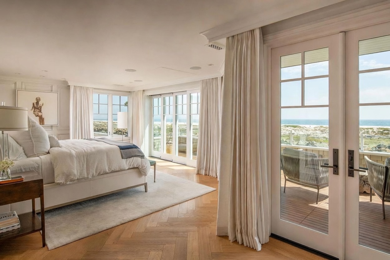 This elegant primary bedroom features light wood herringbone flooring and a neutral color palette that emphasizes a serene, coastal aesthetic. Large glass doors open onto a private deck, providing expansive views of the beach and ocean beyond. The room is furnished with a plush bed, a side table, and soft drapery, creating a bright and airy atmosphere.