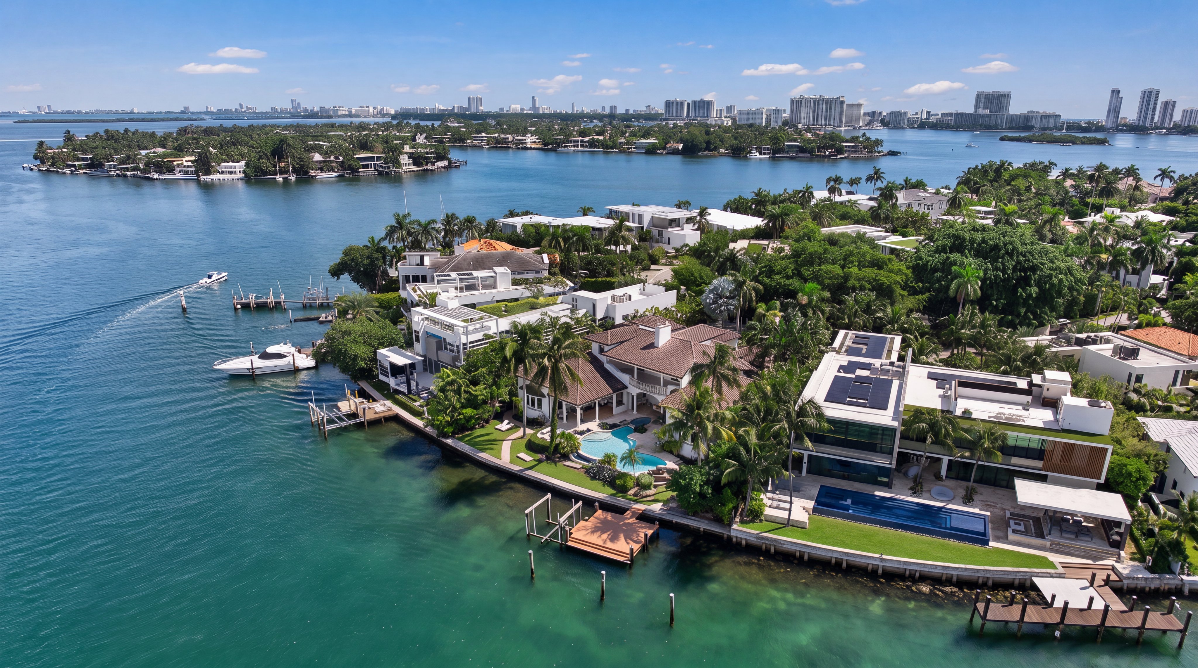 This stunning aerial view highlights a series of luxury waterfront homes situated along a serene bay with clear teal water. The properties feature expansive designs, private docks, multiple swimming pools, and manicured landscaping, reflecting a high-end coastal lifestyle. In the background, the distant city skyline enhances the prestige and prime location of this unique real estate setting.
