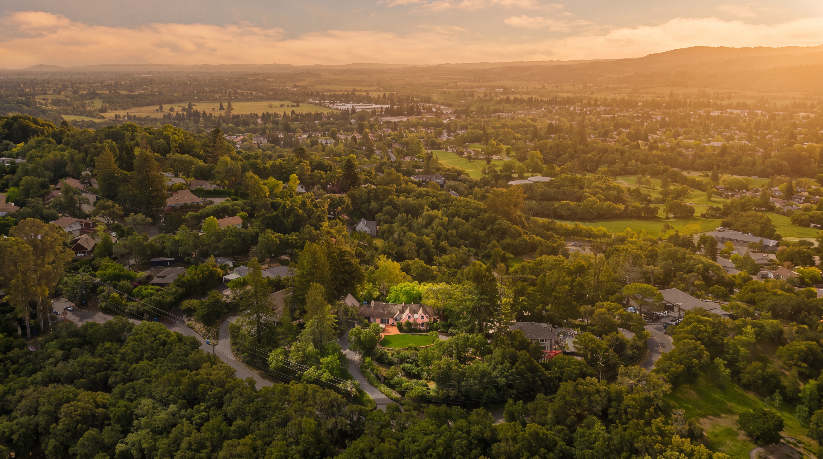 This high-angle aerial perspective captures a secluded residential property nestled within a lush, tree-covered landscape. The home features a traditional architectural style with a multi-gabled roofline, surrounded by mature greenery and a manicured lawn. The expansive view extends across the surrounding valley, showcasing a mix of suburban development and open natural terrain under a bright, partly cloudy sky.
