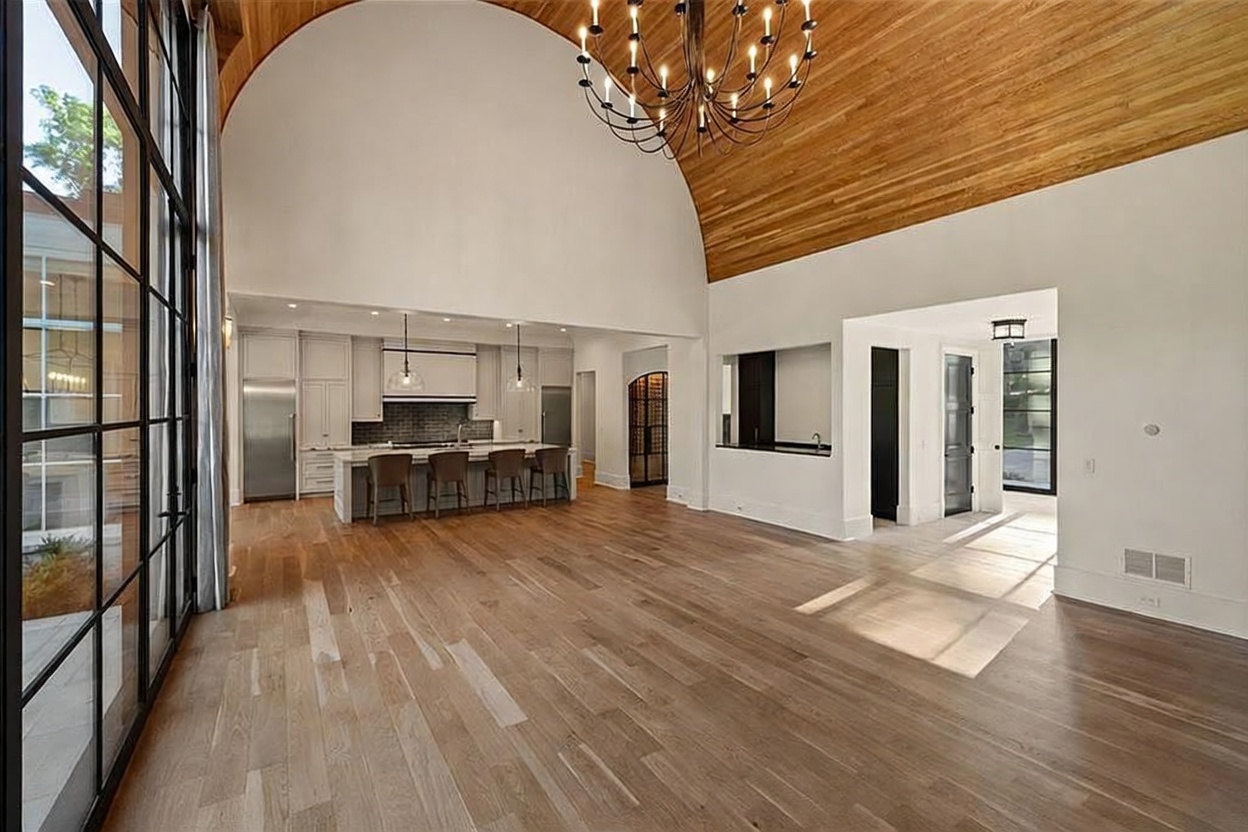 This expansive, open-concept living space features a high, vaulted wood-paneled ceiling and light-toned hardwood flooring throughout. The kitchen area is equipped with a large central island, bar seating, and stainless steel appliances, while a wall of tall, black-framed windows provides abundant natural light. The perspective captures the transition from the kitchen into a bright, airy living area, showcasing a sophisticated and modern architectural design.