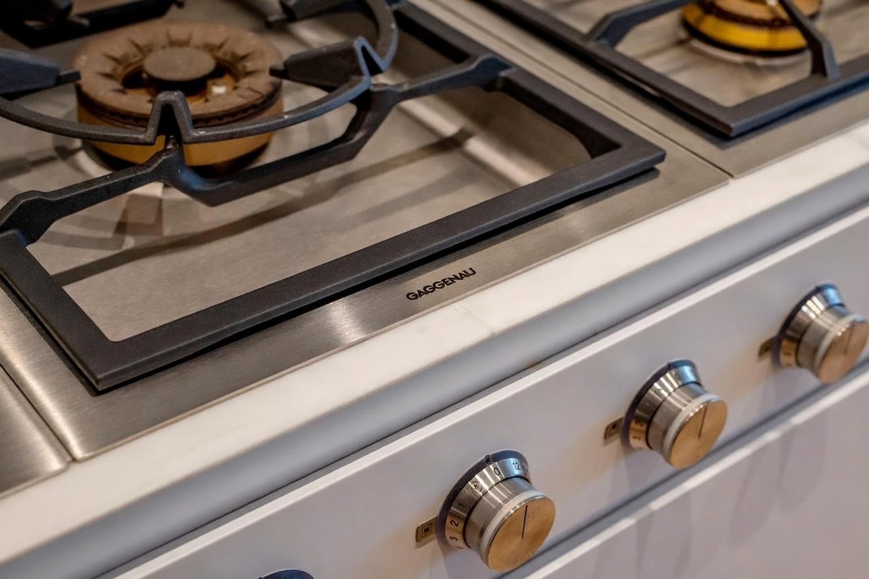 This close-up shot highlights a high-end Gaggenau gas cooktop integrated into a white countertop. The image focuses on the professional-grade burner grates and the sleek, metallic control knobs on the front panel, emphasizing the premium quality of the kitchen appliances. The perspective is angled to showcase the clean lines and modern design of the stove.