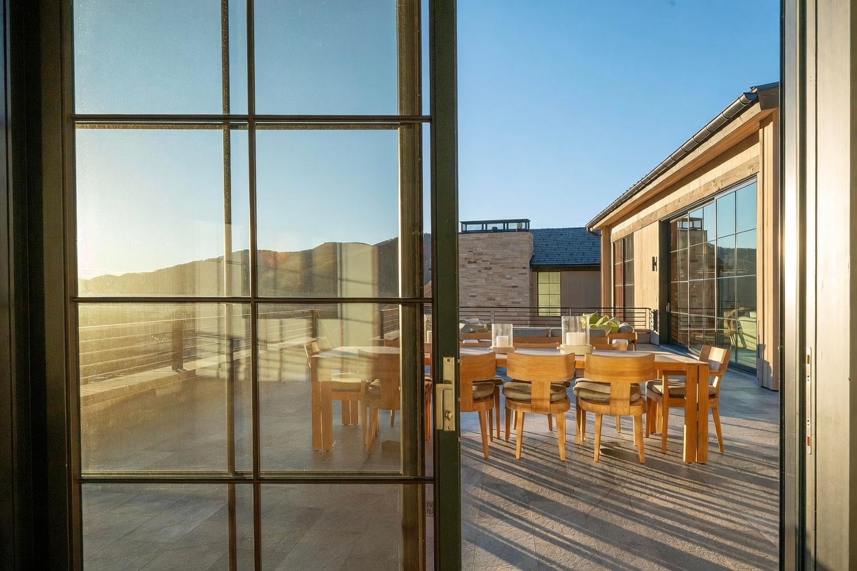 This image captures a spacious outdoor terrace viewed through large, grid-paned glass doors, creating a seamless transition between indoor and outdoor living spaces. The deck is furnished with a long wooden dining table and chairs, set against a backdrop of mountain vistas and a modern building with light-toned siding. The warm, golden-hour sunlight casts long shadows across the stone-tiled floor, emphasizing the expansive and inviting nature of this elevated entertaining area.