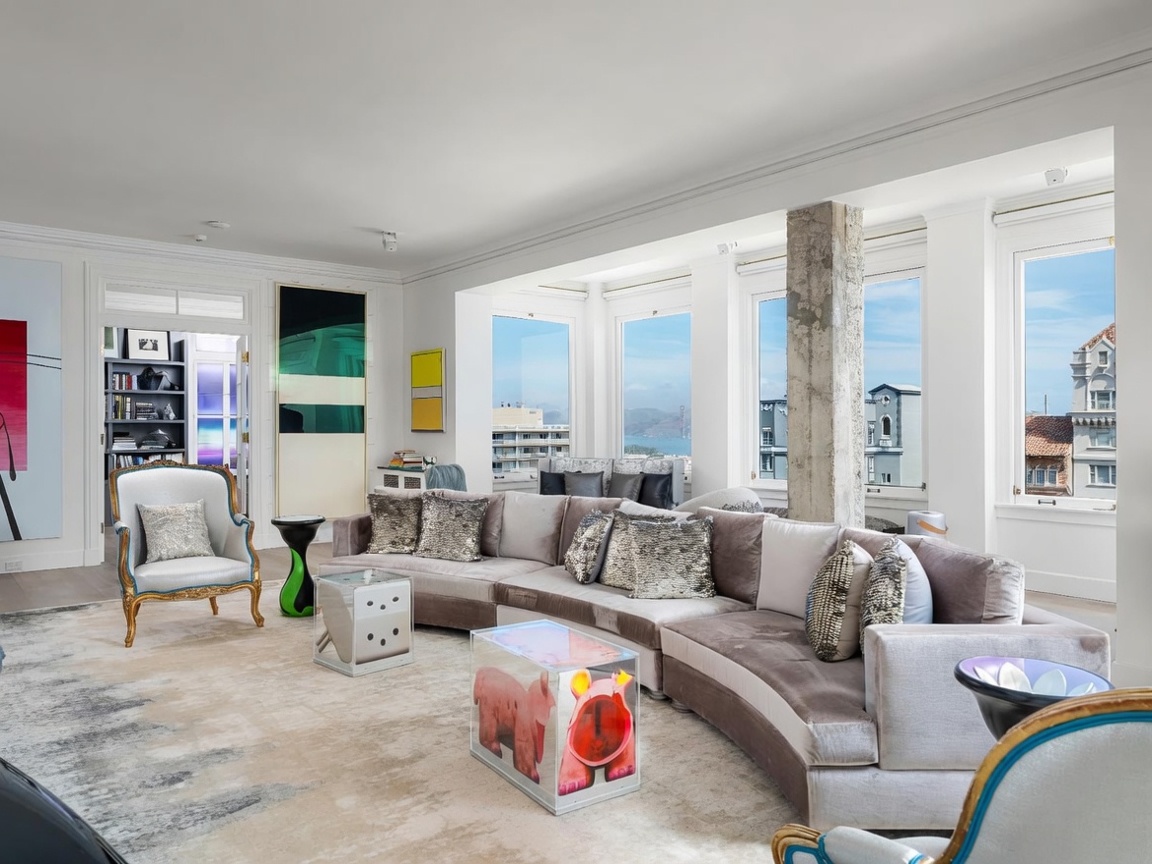 This sophisticated living room features a large, curved sectional sofa upholstered in neutral velvet, accented with metallic throw pillows. The space is anchored by a plush, textured area rug and a unique, transparent coffee table containing a decorative figure. Large windows provide abundant natural light and frame a scenic view of the city and bay, while a prominent concrete column adds an industrial touch to the refined interior.
