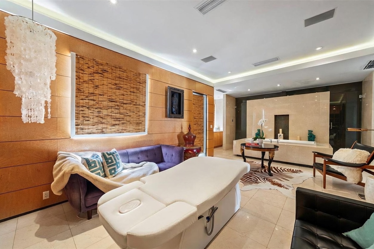 This luxurious spa-inspired room features a central massage table, a plush purple sofa, and a deep soaking tub set against a large stone-clad wall. The space is accented by warm wood-paneled walls, a dramatic chandelier, and a zebra-print rug, creating an atmosphere of high-end relaxation. The perspective is wide-angle, capturing the entire layout from the foreground massage table to the bathing area in the back.