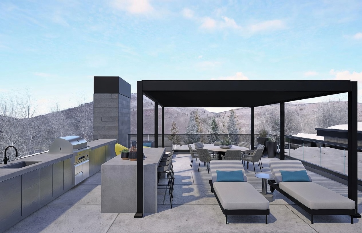 This modern outdoor terrace features a covered dining area with a round table and chairs, adjacent to a sleek outdoor kitchen equipped with a grill and prep counter. A pair of lounge chairs sits on the open deck, which is enclosed by a glass railing that offers unobstructed views of a snowy, mountainous landscape. The space is defined by a contemporary pergola structure and a tall, stone-clad chimney breast, creating a sophisticated environment for outdoor entertaining.