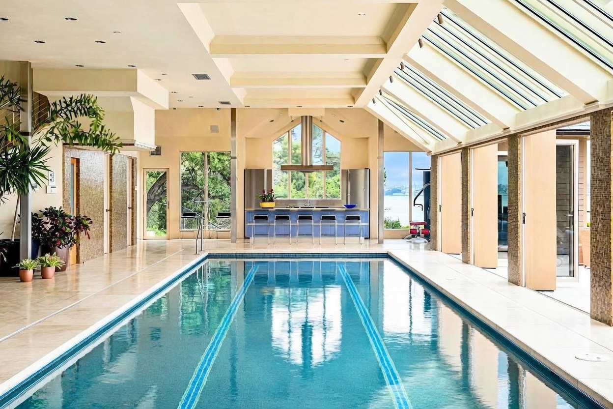 This indoor lap pool area features a long, rectangular pool with dual lane markers, set within a bright, expansive room. The space is defined by a dramatic sloped glass ceiling that floods the area with natural light, complemented by a built-in bar area with stools at the far end. The room includes tiled flooring, neutral-toned walls, and large windows that provide a scenic view of the outdoors, creating a luxurious and serene atmosphere.