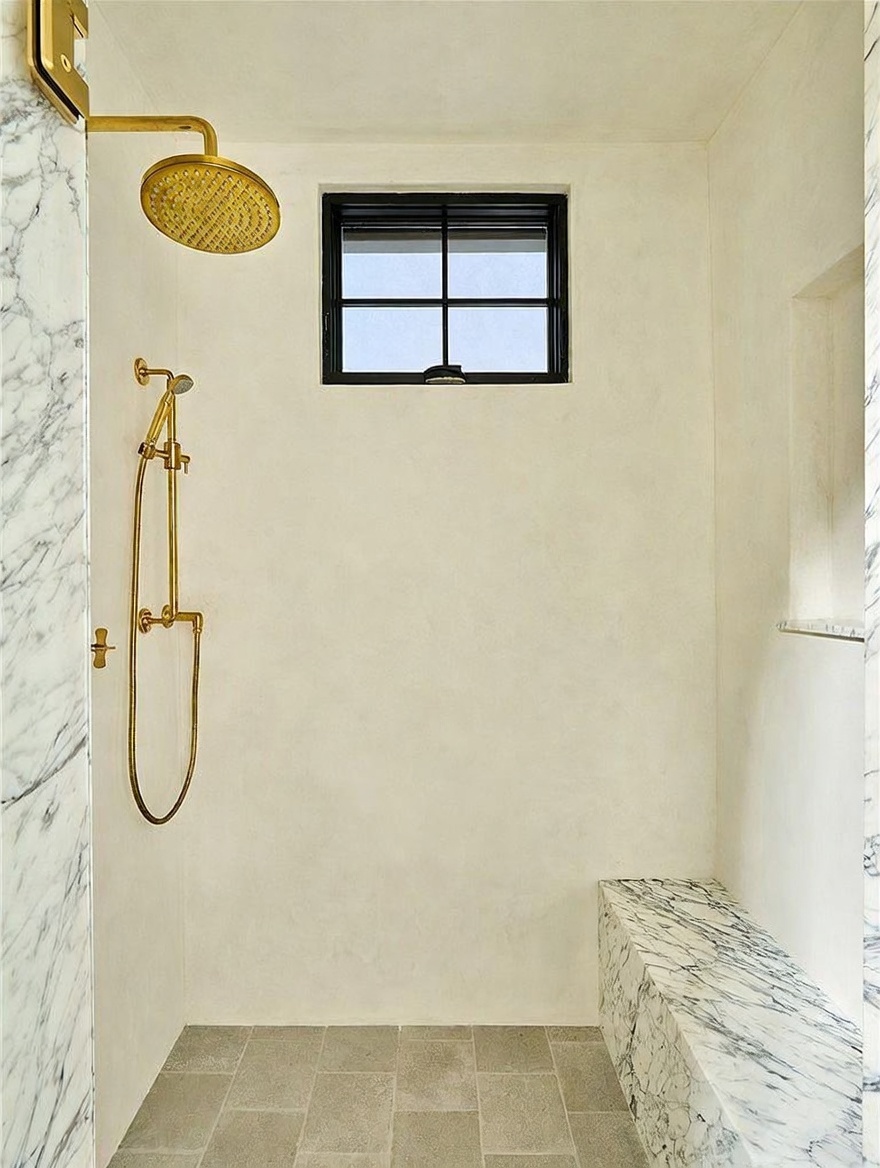 This walk-in shower features a minimalist design with light-toned walls and a stone-tiled floor. A built-in bench with a prominent veined stone finish provides seating, while a gold-toned rainfall showerhead and handheld sprayer add a touch of luxury. A small, grid-paned window allows natural light into the space, creating a bright and clean aesthetic.
