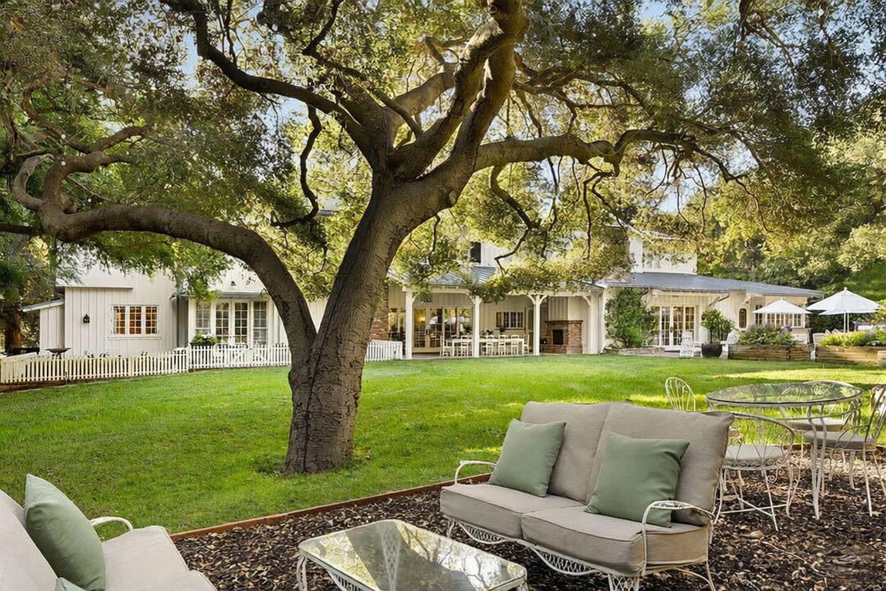 This expansive backyard features a lush green lawn shaded by a large, mature oak tree that serves as a natural canopy. The scene includes a comfortable outdoor seating area with cushioned sofas and a glass-topped table set on a mulch-covered patio, overlooking the white-fenced perimeter of the property. The rear of the white farmhouse-style home is visible in the background, showcasing a covered porch, multiple glass doors, and a brick chimney structure.