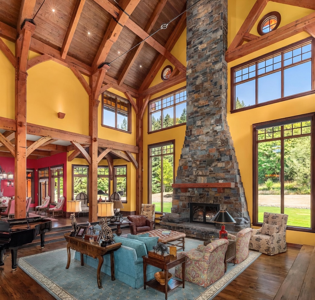 This expansive living room features a dramatic vaulted timber-frame ceiling and a prominent stone-clad chimney breast that serves as the room's focal point. Large windows throughout the space provide abundant natural light and views of the surrounding landscape, while a blue area rug anchors the comfortable seating arrangement. The warm yellow walls and exposed wood beams create a rustic yet grand atmosphere, complemented by a grand piano in the foreground.