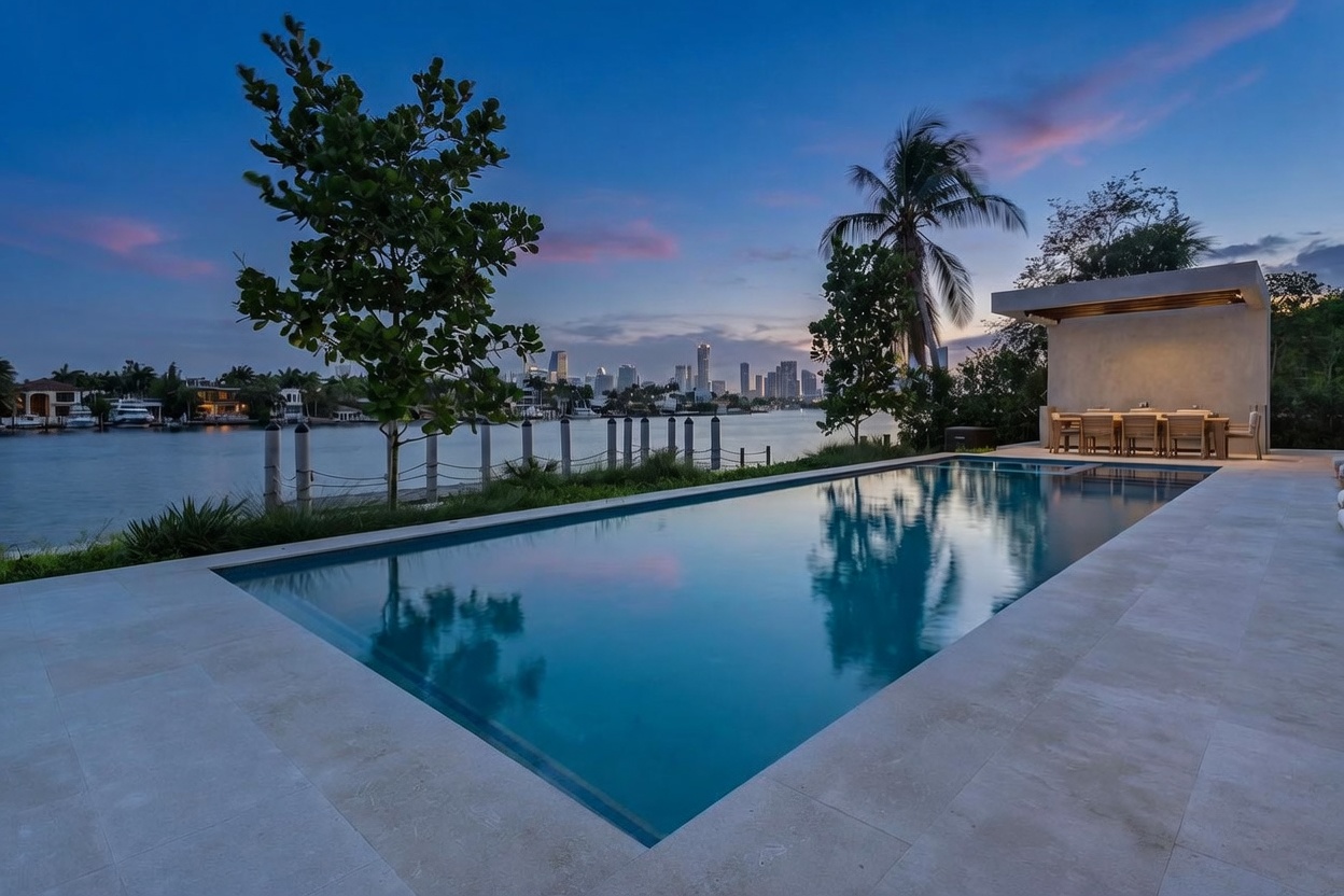 This luxurious waterfront property features a sleek, rectangular infinity-edge pool that mirrors the twilight sky and the distant city skyline. Adjacent to the pool is a covered outdoor dining area with a modern structure, providing a sophisticated space for entertaining. The scene is framed by lush greenery and a palm tree, creating a serene and upscale atmosphere at dusk.