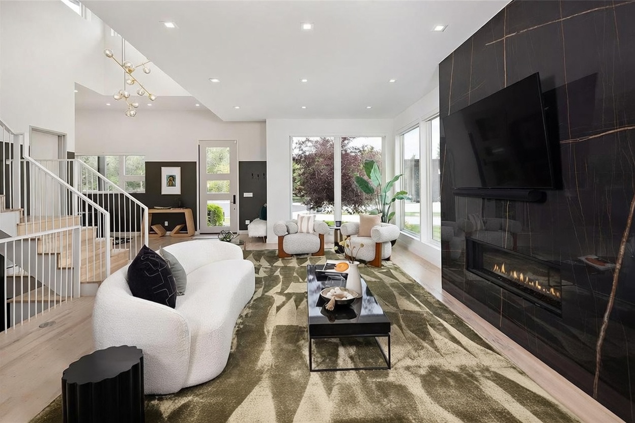 This contemporary living room features a striking black stone-clad wall with an integrated linear fireplace and a large television. The space is furnished with a curved white sofa, two textured armchairs, and a pair of leather accent chairs, all arranged on a large patterned area rug. Large windows provide ample natural light, while a modern chandelier hangs above the adjacent staircase, creating an open and airy atmosphere.