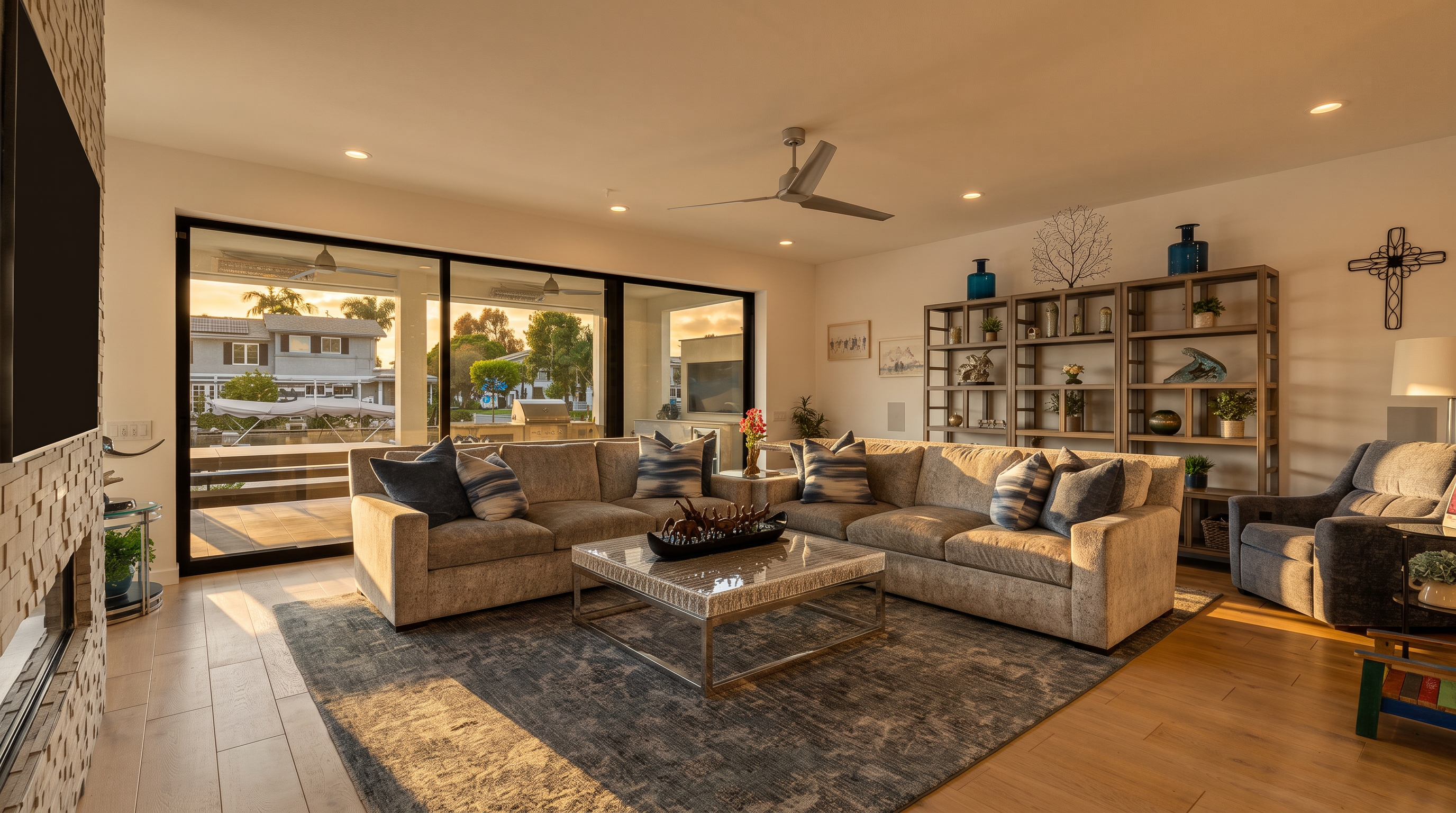 This spacious and contemporary living room features a large, plush sectional sofa centered around a modern coffee table with a metal base. The room is anchored by a textured area rug and includes a tall, open shelving unit filled with decor, alongside a comfortable armchair. Expansive glass sliding doors provide a seamless transition to the outdoor space, filling the room with natural light and offering a view of the surrounding exterior.