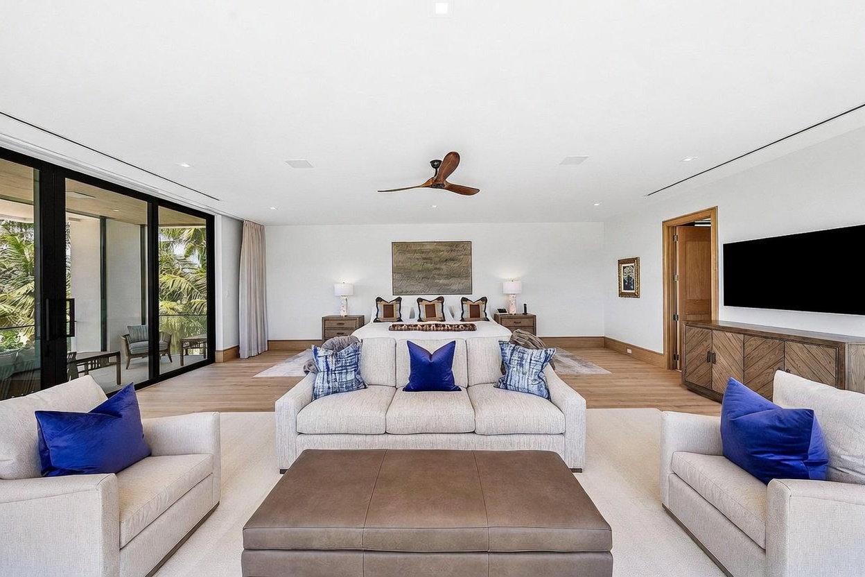 This expansive primary bedroom suite features a sophisticated open-concept layout that seamlessly integrates a lounge area with a king-sized bed. The room is anchored by a large, neutral-toned sofa and a leather ottoman in the foreground, while floor-to-ceiling glass sliding doors provide access to a private balcony with lush tropical views. The space is finished with light wood flooring, a central ceiling fan, and a wall-mounted television, creating a serene and luxurious retreat.