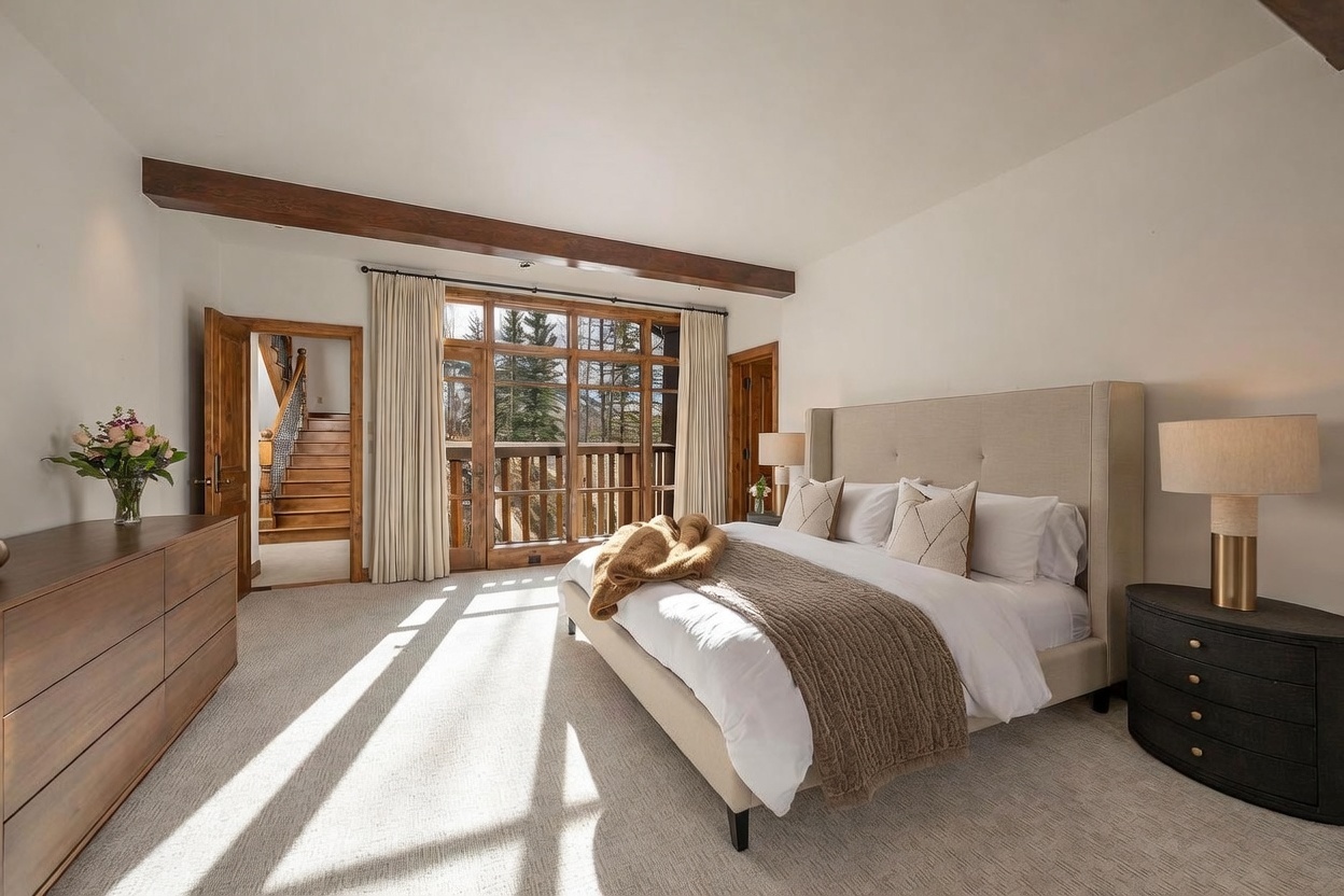 This spacious primary bedroom features a neutral color palette, a large upholstered bed with layered bedding, and a dark wood dresser. A prominent wooden ceiling beam adds architectural character, while large windows and a glass door provide ample natural light and a view of the wooded exterior. The room is accented by two bedside lamps and a vase of flowers, creating a serene and inviting atmosphere.