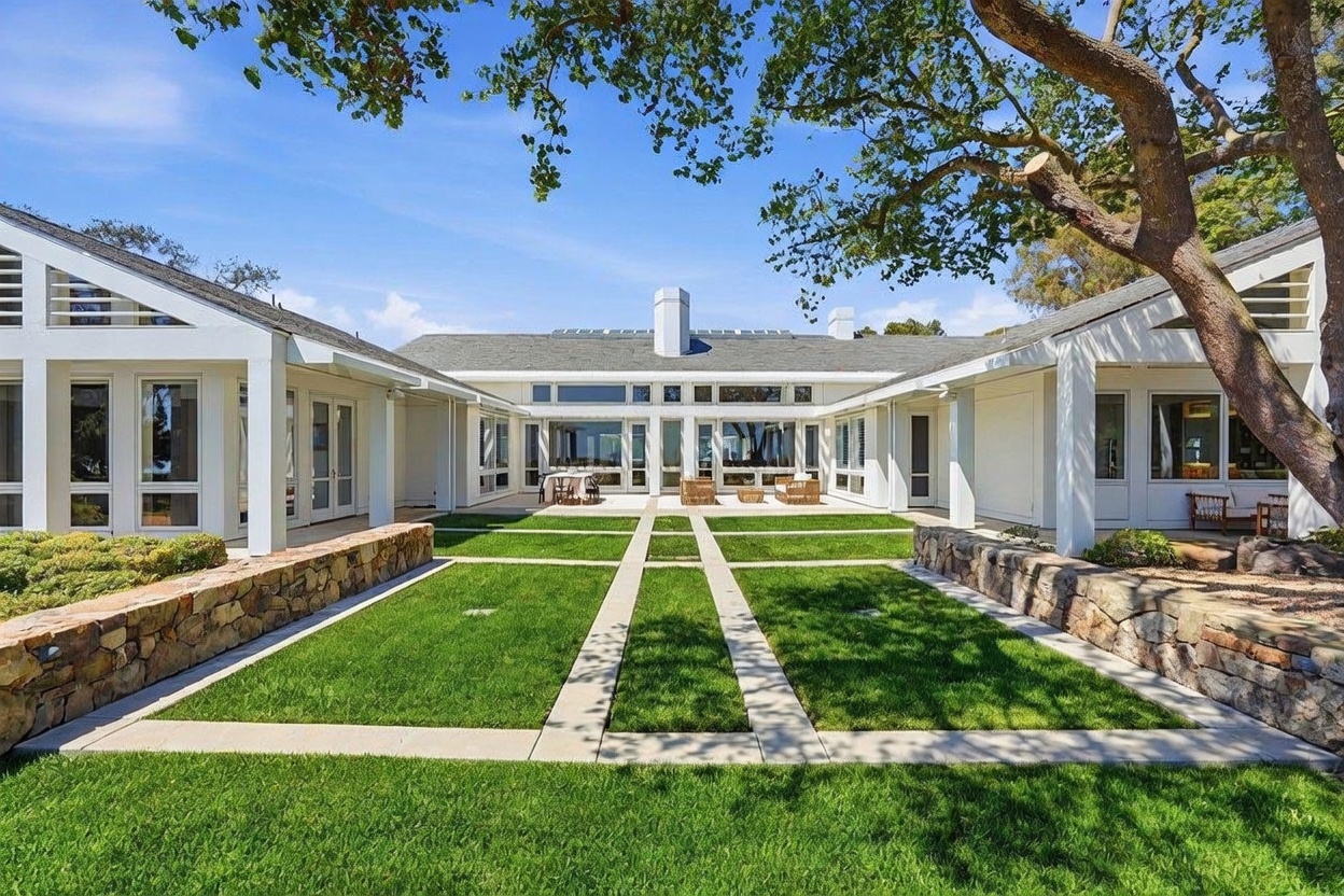 This rear view of a sprawling, single-story residence showcases a U-shaped architectural design centered around a meticulously manicured courtyard. The home features white siding, large windows, and a classic shingled roof, with a stone-lined garden path leading toward the central patio area. A large, mature tree frames the right side of the composition, casting dappled shade over the lush green lawn and stone retaining walls.