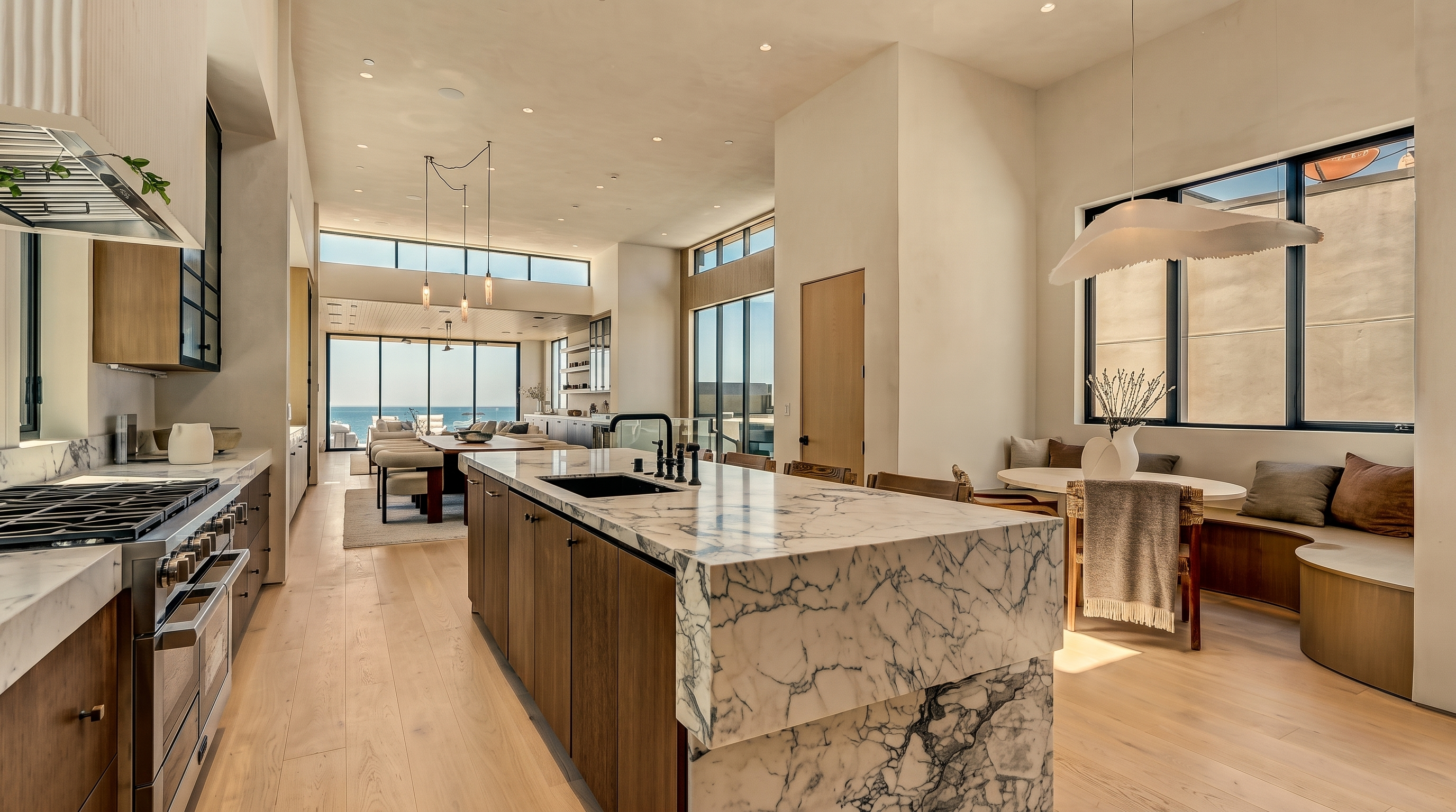 This expansive, modern kitchen features a large central island with a waterfall-edge stone countertop and integrated sink. The space is characterized by light wood cabinetry, high ceilings, and a seamless transition into an adjacent dining area with built-in bench seating. Large windows provide abundant natural light and a clear view of the ocean in the distance, creating an airy and sophisticated atmosphere.
