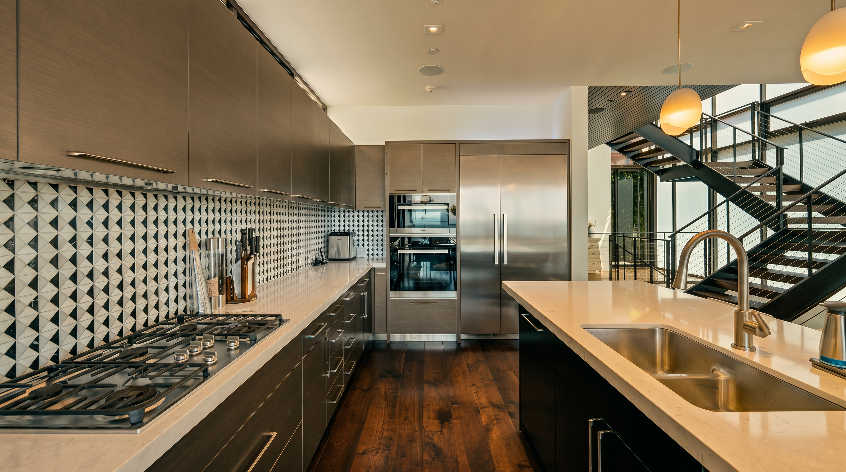 This modern kitchen features sleek, dark-toned cabinetry paired with light-colored stone countertops and a striking geometric patterned tile backsplash. A large central island provides additional workspace and includes a stainless steel sink, while a professional-grade gas range and built-in stainless steel appliances complete the layout. The space is open to a staircase with a metal railing, creating a contemporary and airy atmosphere with rich wood flooring throughout.