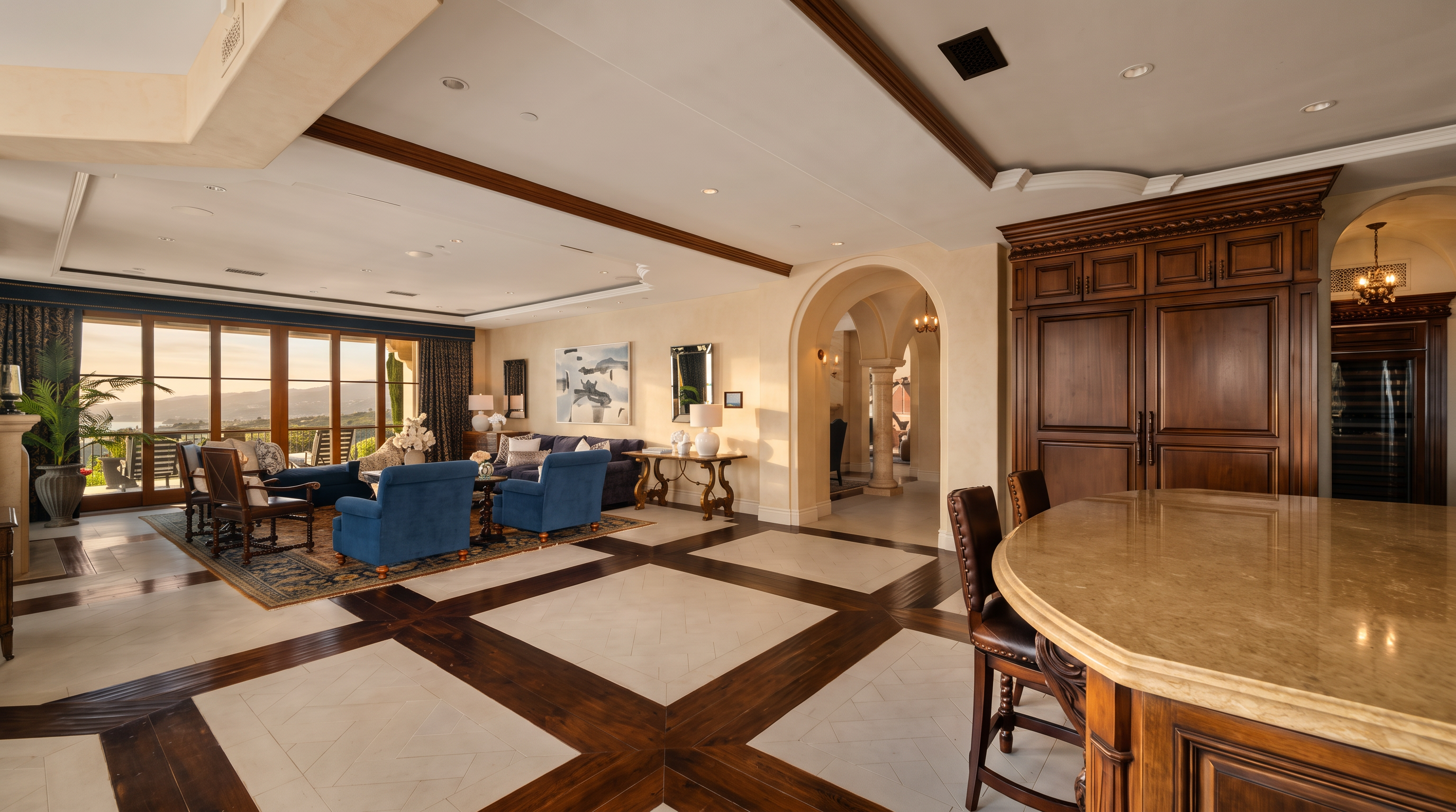 This expansive open-concept living area features a sophisticated blend of dark wood and light stone flooring arranged in a geometric pattern. The space includes a comfortable seating area with blue armchairs and a sofa, positioned near large windows that provide a view of the outdoors. A large kitchen island with a stone countertop occupies the foreground, while an arched doorway leads to an adjacent room, creating a sense of depth and architectural elegance.