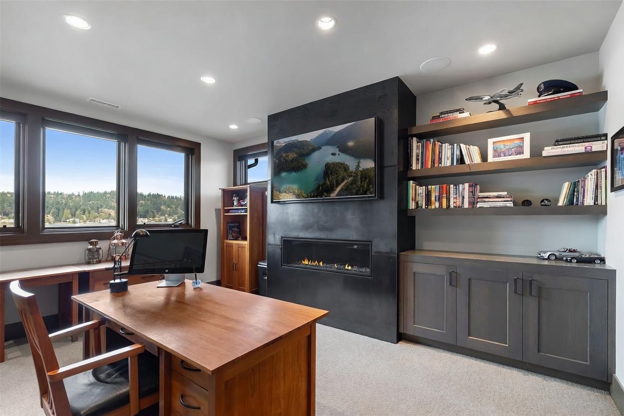 This sophisticated home office features a large wooden desk positioned to face a dark, full-height fireplace surround with an integrated television. To the right, built-in shelving and cabinetry provide ample storage, while large windows on the left offer a scenic view of the surrounding landscape. The room is finished with neutral carpeting and recessed lighting, creating a professional and tranquil workspace.