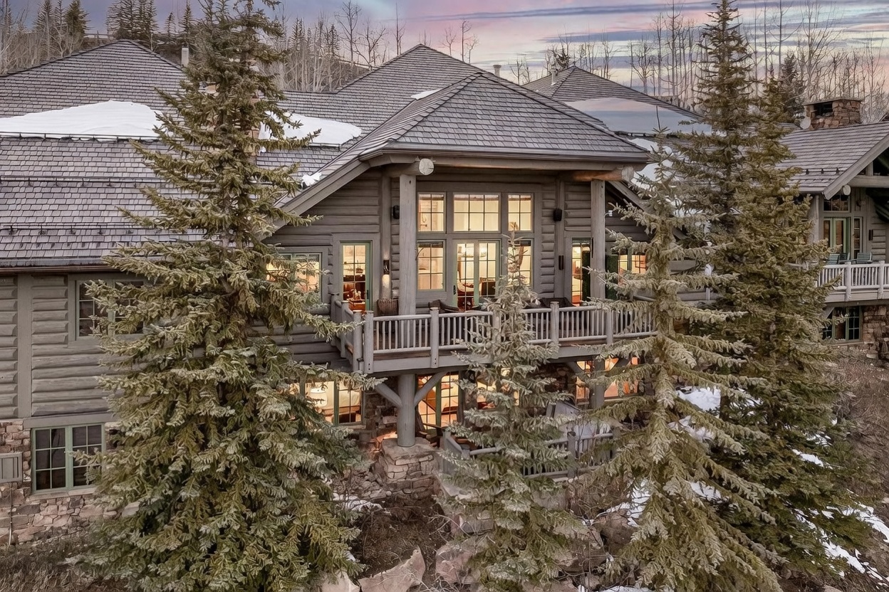 This impressive mountain estate features a rustic log construction with a multi-level design and a dark shingled roof. The rear facade showcases a prominent wooden deck and a lower-level patio, both offering views of the surrounding evergreen forest and rocky terrain. Large windows and glass doors allow natural light to fill the interior spaces, creating a warm and inviting atmosphere against the backdrop of a twilight sky.