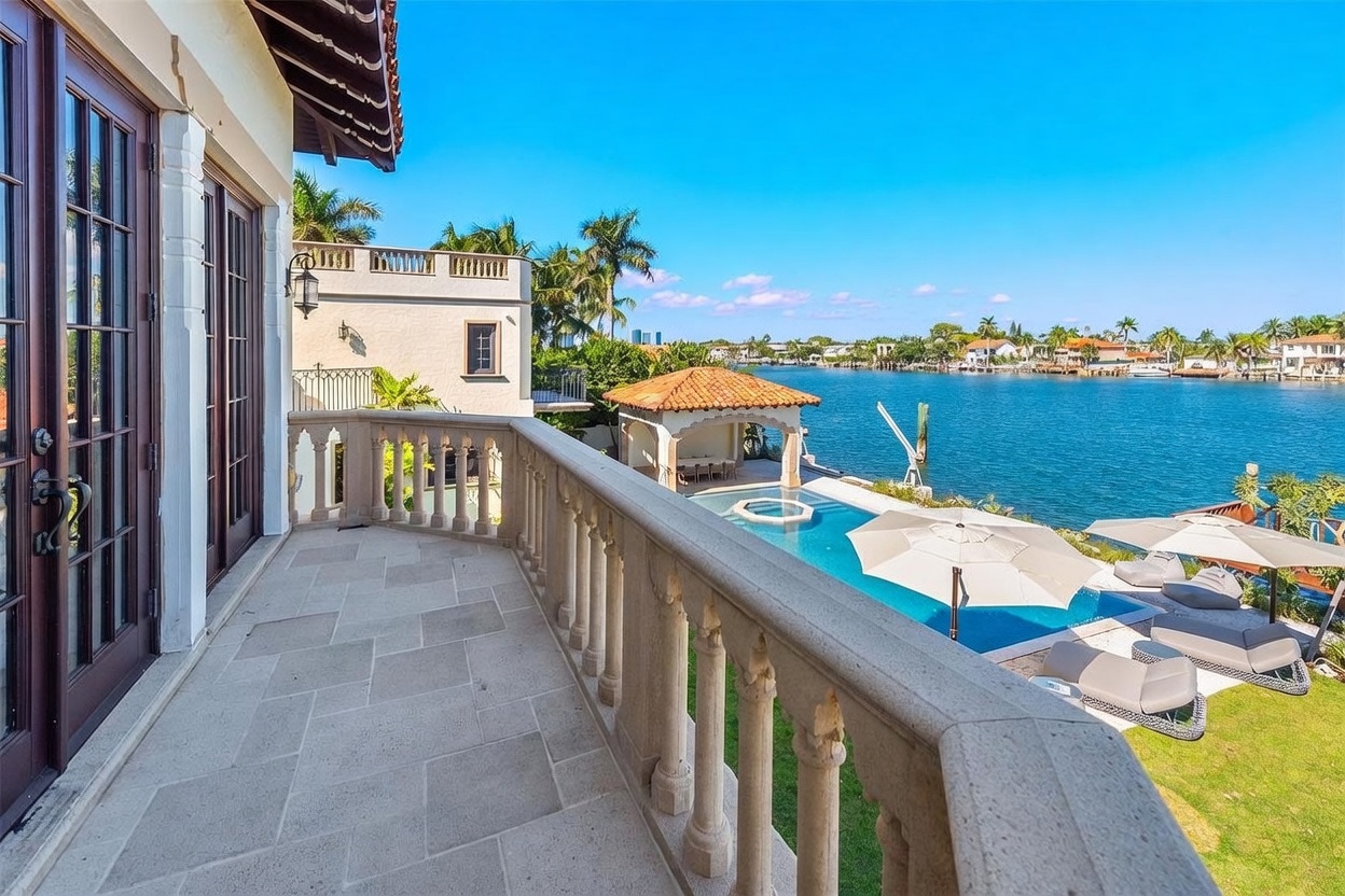 This elevated stone balcony offers a panoramic view of a waterfront property, featuring a private pool, a covered cabana, and a lush green lawn. The balcony is enclosed by a classic stone balustrade and provides direct access to the home through large glass doors. The scene captures a serene, high-end coastal lifestyle with clear blue skies and sparkling water.