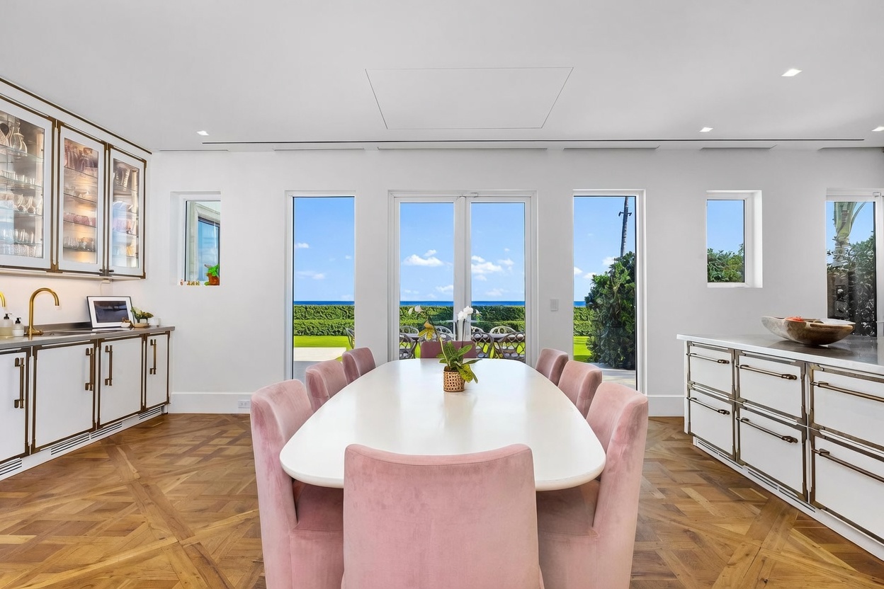 This elegant dining area features a long, white oval table surrounded by soft pink upholstered chairs, set against a backdrop of large glass doors that provide a clear view of the ocean. The room is anchored by a sophisticated herringbone-patterned wood floor and is flanked by white cabinetry with metallic trim on both sides. The bright, airy atmosphere is enhanced by the natural light streaming in, creating a serene and luxurious coastal dining experience.