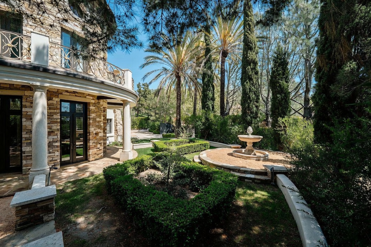 This elegant backyard garden features a stone-clad home exterior with a curved patio and a decorative wrought-iron balcony railing. A central stone fountain serves as a focal point, surrounded by manicured hedges and tall, slender cypress trees that create a Mediterranean-inspired atmosphere. The perspective captures the transition from the home's stone architecture to the lush, sun-drenched landscaping.