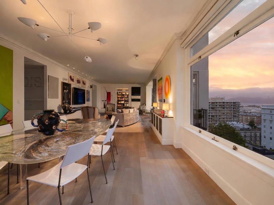 This sophisticated open-concept living and dining area features light wood flooring and a striking, modern multi-arm chandelier. A unique, organic-shaped dining table serves as a focal point in the foreground, while the living area beyond includes a curved sofa and a wall-mounted television. Large windows along the right wall provide an expansive, elevated view of the city skyline and bay at sunset, creating a bright and airy atmosphere.