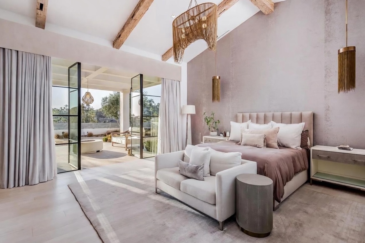 This luxurious primary bedroom features a vaulted ceiling with exposed wooden beams and a soft, neutral color palette. A plush sofa sits at the foot of the bed, while large glass doors open directly onto a sun-drenched patio, creating a seamless indoor-outdoor living experience. The room is accented by unique, tiered fringe pendant lights and a textured feature wall, offering a sophisticated and tranquil atmosphere.