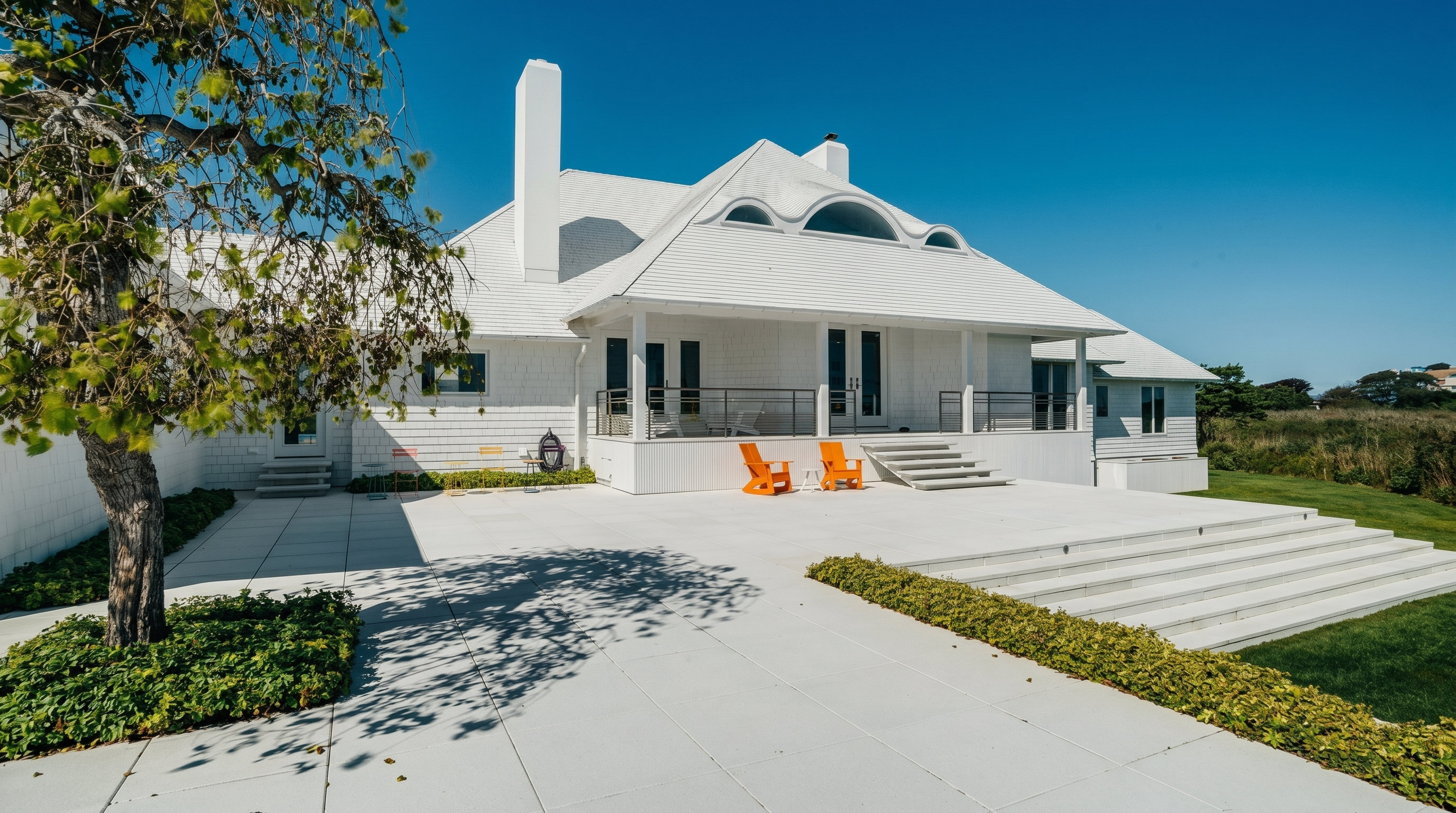 This rear view of a coastal-style home showcases a expansive light-colored stone patio leading to a raised deck with metal railings. The house features white shingle siding, a prominent chimney, and a roofline accented by arched dormer windows. A large, mature tree provides shade on the left, while the open lawn and clear blue sky create a bright, airy, and inviting atmosphere.