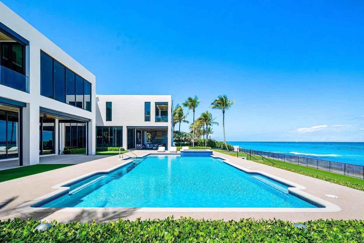 This stunning modern estate features a large, rectangular swimming pool with an integrated spa, set against a backdrop of the deep blue ocean. The home showcases clean, white architectural lines with expansive glass walls that seamlessly connect the indoor and outdoor living spaces. A manicured lawn and lush green hedges frame the pool deck, creating a serene and luxurious coastal retreat.