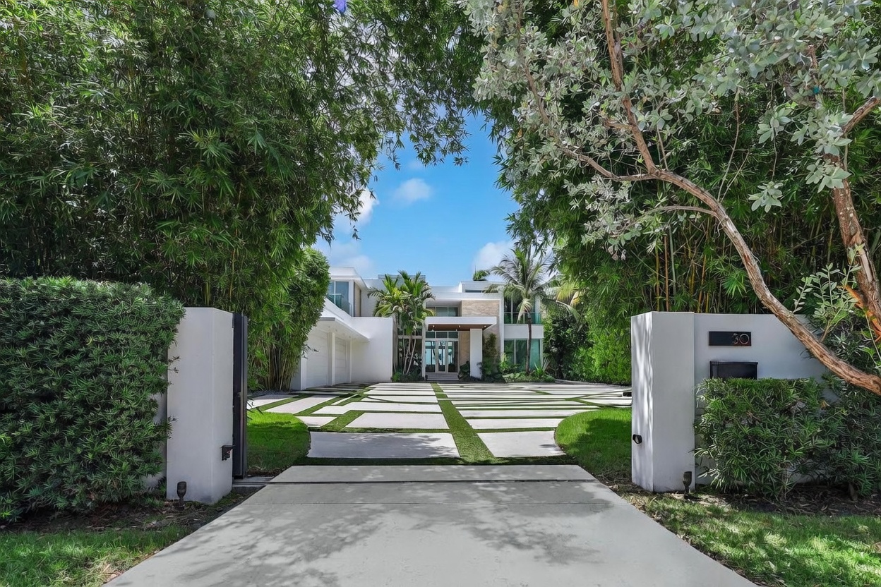 This modern luxury residence features a striking contemporary design with clean lines and a prominent white facade. The property is accessed via a wide, geometric paver driveway framed by lush, tall bamboo hedges that create a private, resort-like atmosphere. The perspective is a straight-on, eye-level shot from the gated entrance, highlighting the grand scale of the home and its manicured landscaping.