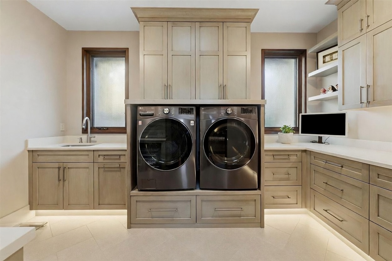 This well-appointed laundry room features custom light-toned cabinetry surrounding a pair of dark-finished front-load washer and dryer units. The space includes a built-in sink, ample counter space with a light-colored surface, and under-cabinet lighting that creates a bright, functional atmosphere. Two windows with textured glass provide natural light, while the neutral color palette and clean lines offer a modern and organized aesthetic.