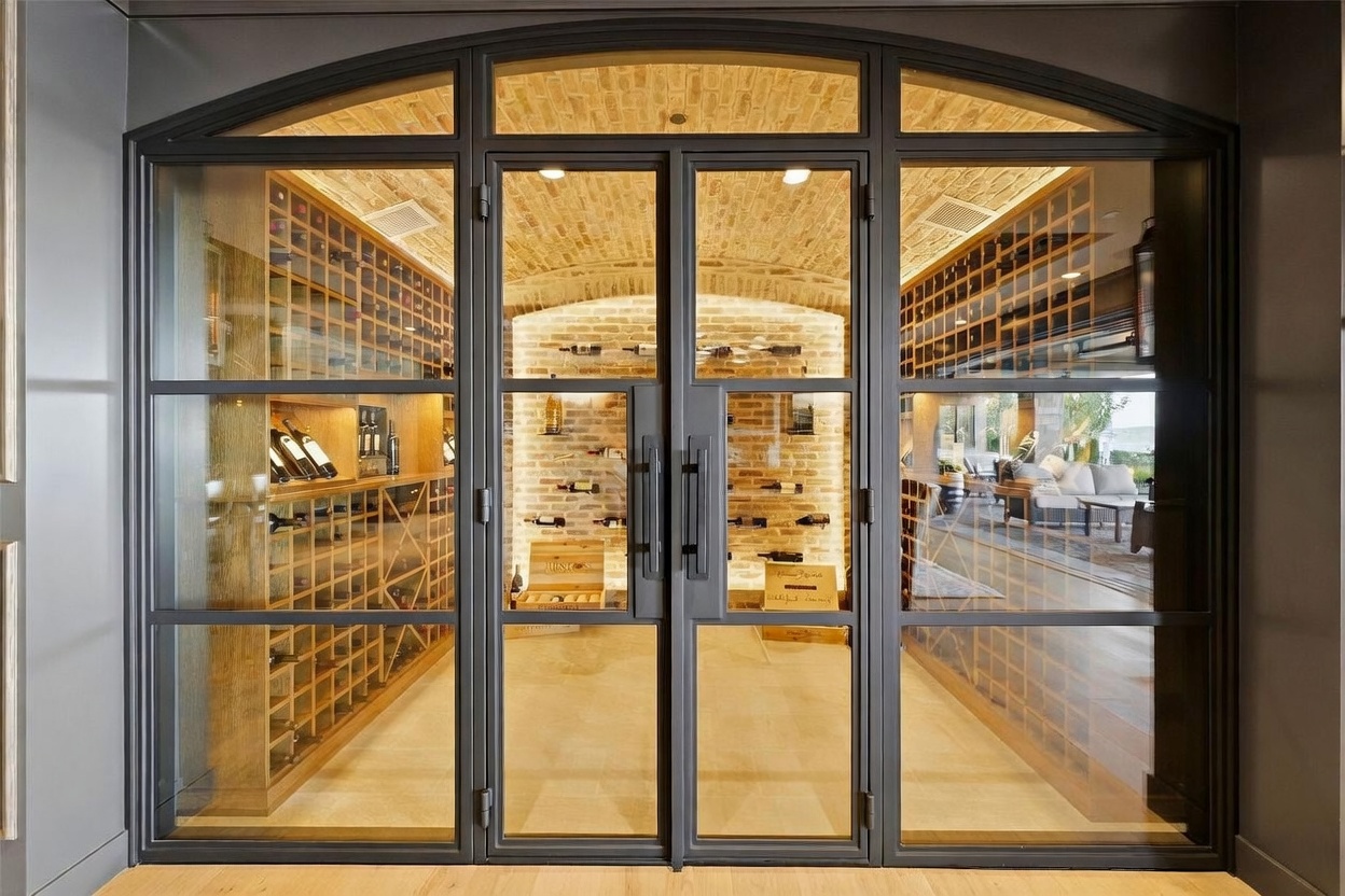 This sophisticated wine cellar features a striking arched iron-framed glass door entry that provides a clear view into the climate-controlled space. Inside, the room is lined with custom light wood wine racking and showcases a rustic brick barrel-vaulted ceiling. The combination of warm ambient lighting and the textured brick backdrop creates an elegant, high-end atmosphere for wine storage.