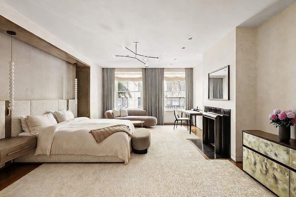 This spacious primary bedroom features a neutral, sophisticated palette with a large bed centered against a textured wall panel. The room includes a curved sofa near the windows, a dedicated desk area, and a dark stone fireplace surround that adds a dramatic focal point. Large windows with floor-length curtains allow ample natural light to fill the room, creating an airy and luxurious atmosphere.