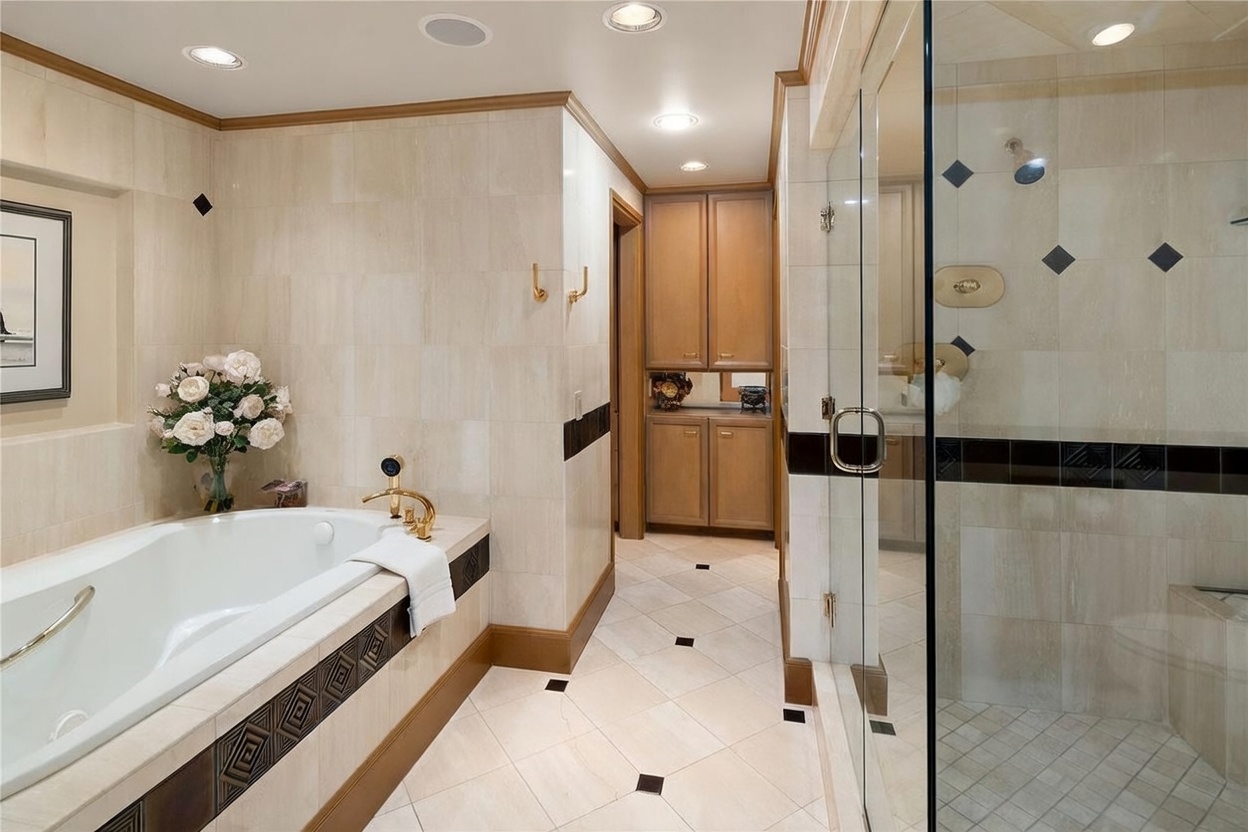 This primary bathroom features a deep soaking tub with gold-toned fixtures and a separate glass-enclosed shower. The space is finished with light-toned wall tiles accented by a dark horizontal border, complemented by light wood cabinetry and patterned floor tiles. The perspective captures the transition from the bathing area toward a small vanity nook at the end of the hallway.